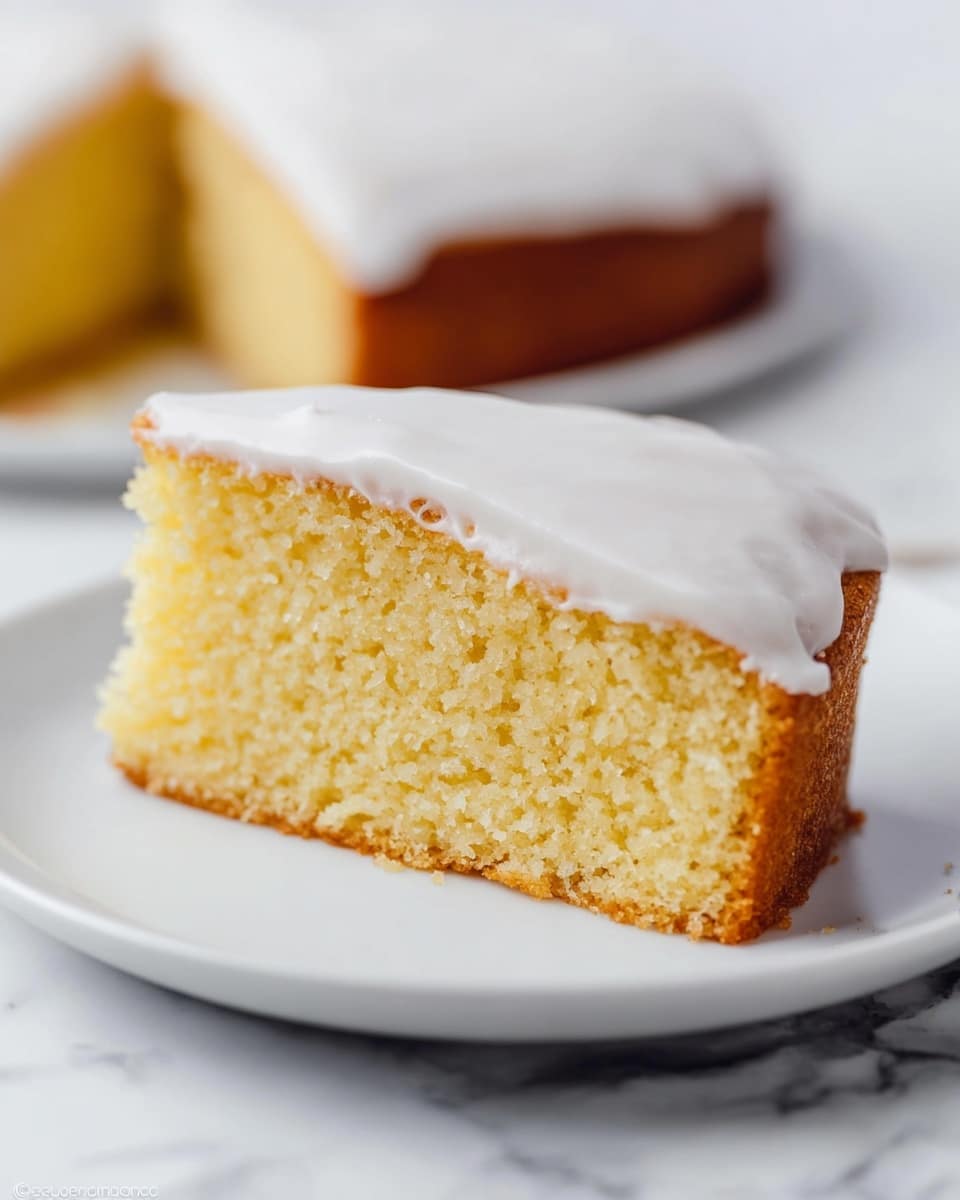 A single slice of light yellow cake with a soft, fluffy texture and a light brown crust sits on a white plate. The cake has one visible layer, topped with a smooth, thick white icing that covers the entire top surface. The background shows the rest of the cake slightly blurred, also covered with the same white icing. The scene is set on a white marbled surface. photo taken with an iphone --ar 4:5 --v 7