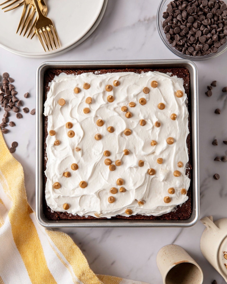 A square dessert with two visible layers in a metal baking tray on a white marbled surface: the bottom layer is a dark brown, dense-looking cake, and the top layer is a thick, fluffy white cream spread unevenly but fully covering the cake. Scattered on the cream are small light brown and dark brown chocolate chips. Around the tray, there is a white plate with two forks, a yellow and white striped cloth, a glass bowl of dark brown chocolate chips, and a ceramic container. Photo taken with an iphone --ar 4:5 --v 7