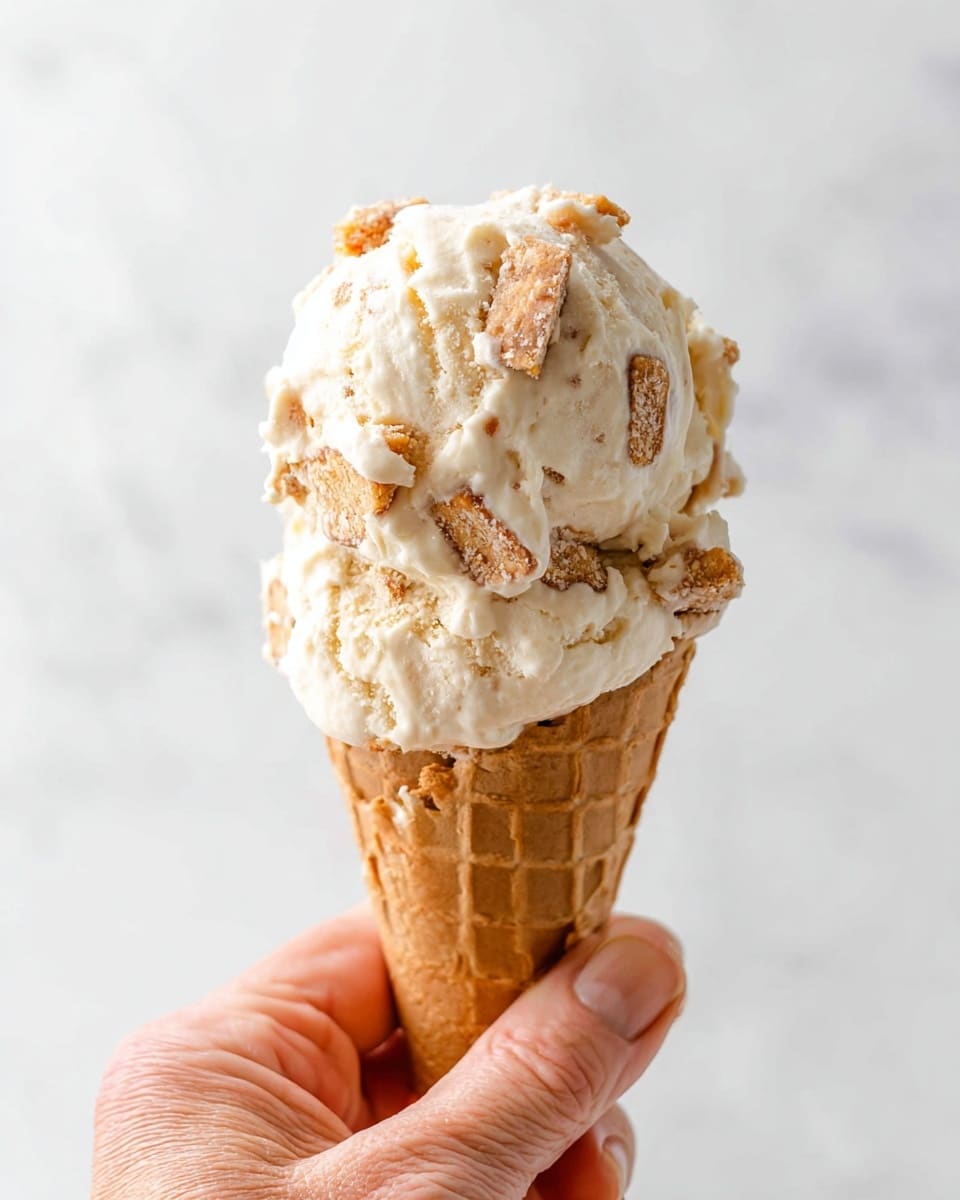 A close-up image of a single scoop of light beige ice cream with visible chunks of a golden-brown biscuit mixed inside, sitting atop a light brown waffle cone with a classic crisscross pattern. The cone is held by a woman's hand with slightly wrinkled skin, visible fingernails, and a relaxed grip. The background and surface beneath the cone have a smooth white marbled texture creating a clean and bright setting. photo taken with an iphone --ar 4:5 --v 7