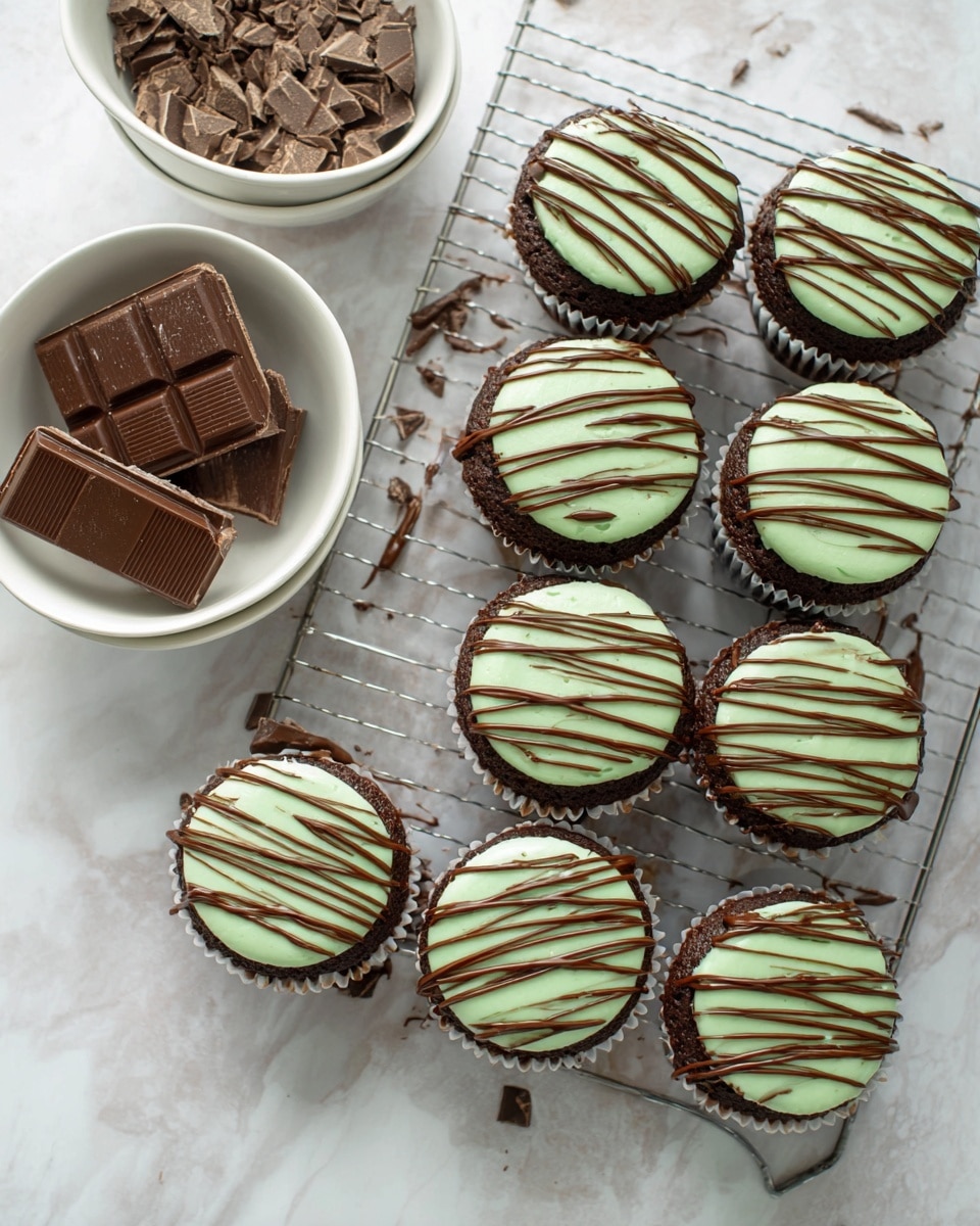 There are eight chocolate cupcakes arranged on a cooling rack over a white marbled surface. Each cupcake has two layers: the bottom layer is dark brown chocolate cake, topped with a smooth pale green frosting layer. All cupcakes have a drizzle of milk chocolate sauce in diagonal lines across the top. To the left of the cupcakes, there are two white bowls on the same white marbled surface; one bowl contains broken pieces of mint chocolate, and the other bowl contains whole mint chocolate chocolate bars stacked inside. Photo taken with an iphone --ar 4:5 --v 7