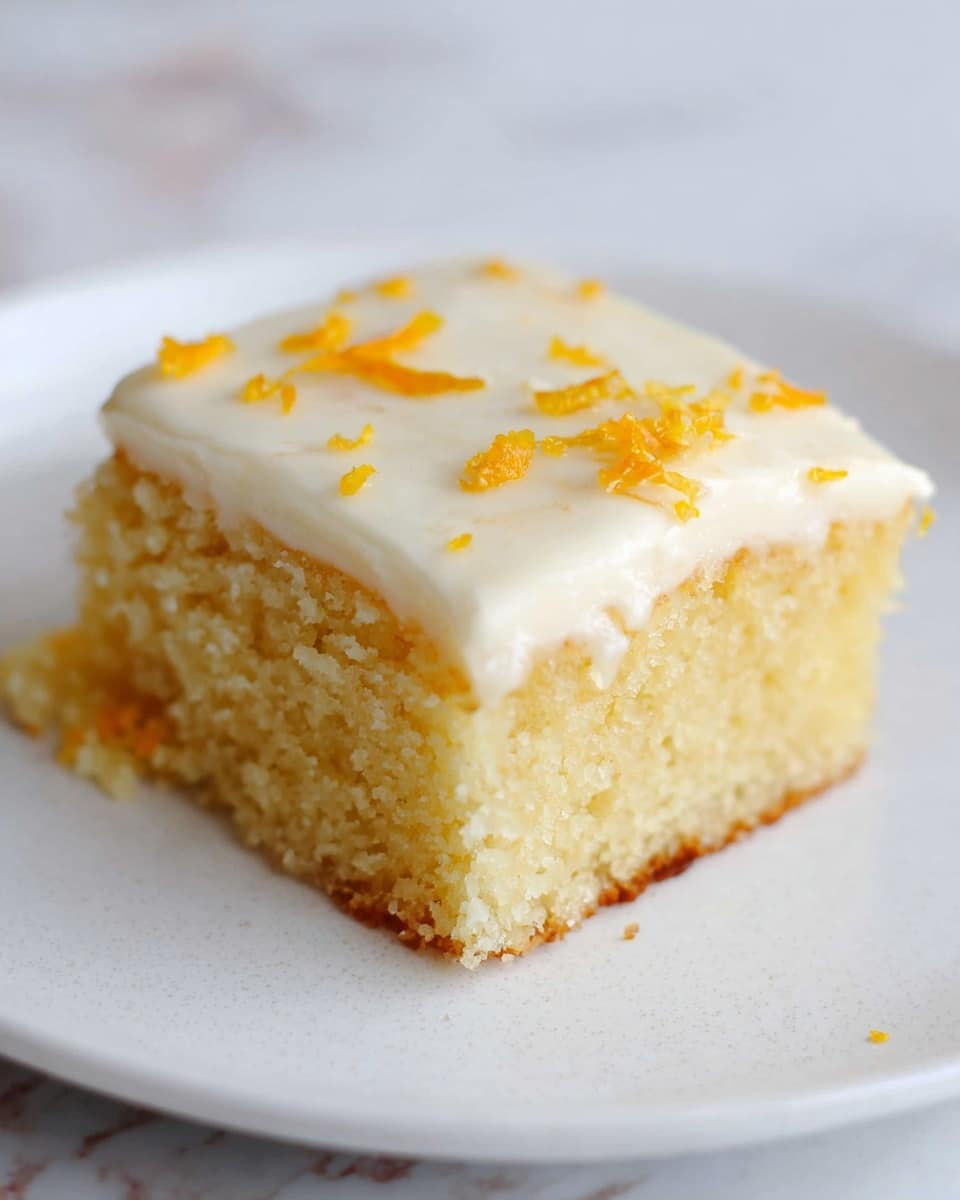 A single square piece of light yellow cake with a soft, crumbly texture sits centered on a white plate. The cake has one distinct layer topped with a smooth, creamy white frosting. Scattered small orange zest pieces are sprinkled on the frosting’s surface, adding a pop of color. The plate rests on a white marbled texture background. photo taken with an iphone --ar 4:5 --v 7