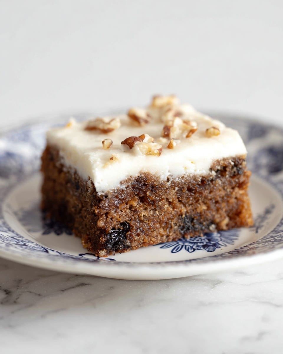 A square piece of cake sits on a white plate with blue flower patterns, placed on a white marbled surface. The cake has two layers: a bottom thick brown layer with a rough texture filled with dark bits and small nut pieces, and a top creamy white layer that looks smooth and soft with some small chopped nuts sprinkled on top. The edges of the cake are sharp and clean, showing a clear separation between the brown base and white frosting photo taken with an iphone --ar 4:5 --v 7