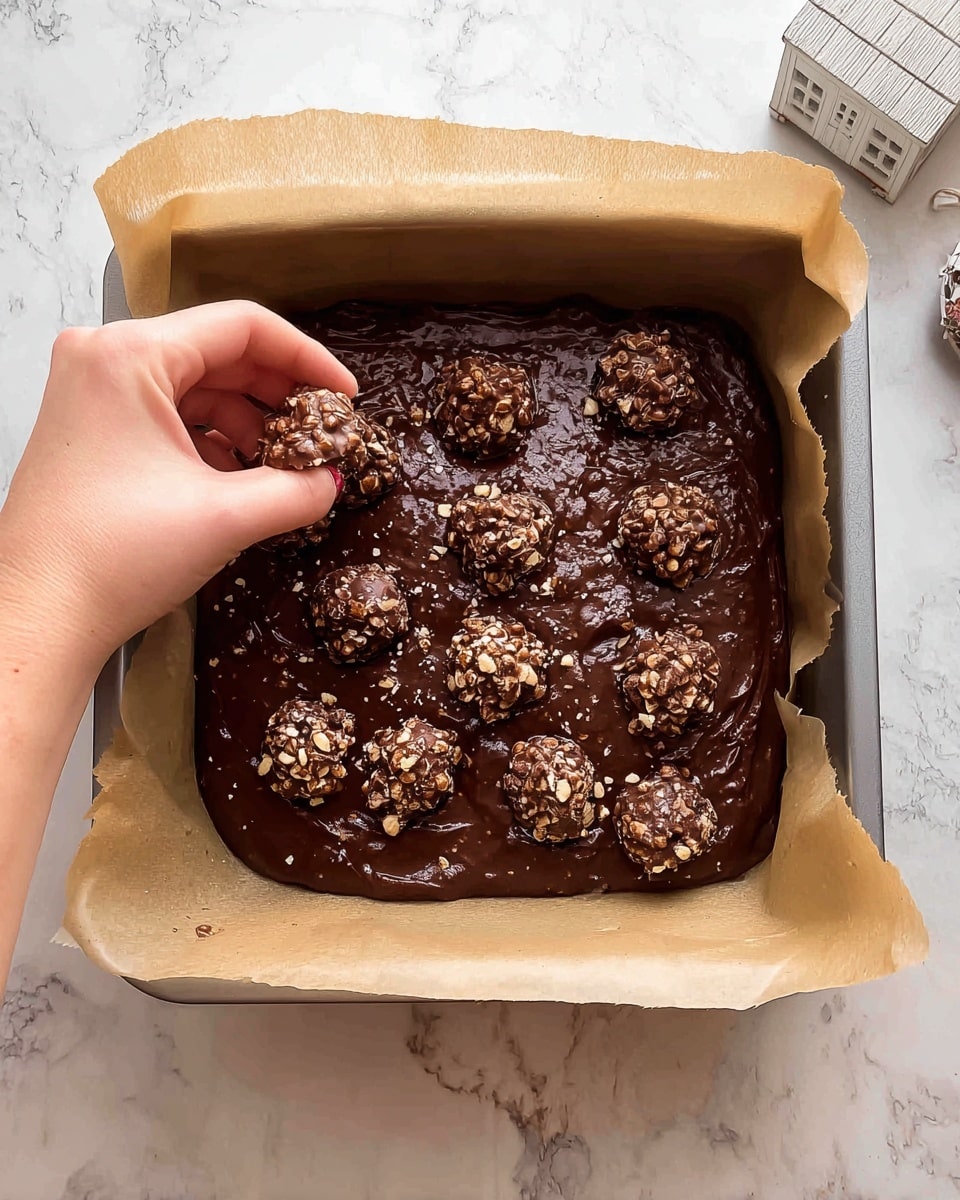 A square metal baking pan lined with brown parchment paper holds a thick, dark brown chocolate batter spread evenly inside. On top of the batter, several whole and broken round chocolate pieces with a nutty, textured surface are scattered, adding a rough, crunchy look. A woman's hand is seen gently placing a piece of the chocolate nut clusters on the batter. The pan rests on a white marbled surface with a small white house-shaped ornament nearby. Photo taken with an iphone --ar 4:5 --v 7