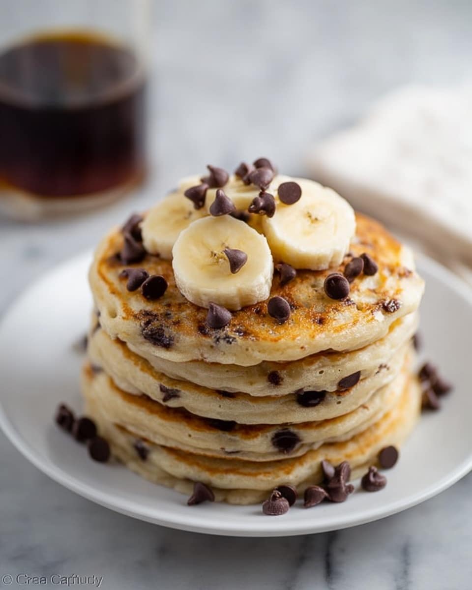 A stack of five round pancakes with a light brown color and chocolate chips baked inside sits on a white plate placed on a white marbled surface. The top pancake is decorated with five banana slices arranged in a small circle, each topped with numerous small dark brown chocolate chips scattered over and around the banana. In the blurred background, there is a glass container with dark liquid inside. The photo taken with an iphone --ar 4:5 --v 7