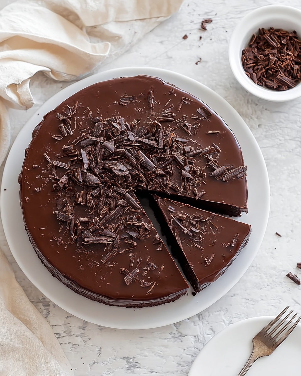 A round chocolate cake with a smooth, shiny dark brown chocolate glaze covering the top and sides sits on a white plate. The top is decorated with dark chocolate shavings scattered mostly around the edges, creating a rough texture contrast with the glossy glaze. One slice is cut but still in place, showing the thickness of the cake which appears dense and moist. The plate is placed on a white marbled textured surface, and a cream-colored cloth is draped casually on the left side of the cake. In the background, a small white bowl holds more chocolate shavings, and a white plate with a fork lies at the bottom right corner of the image. Photo taken with an iphone --ar 4:5 --v 7