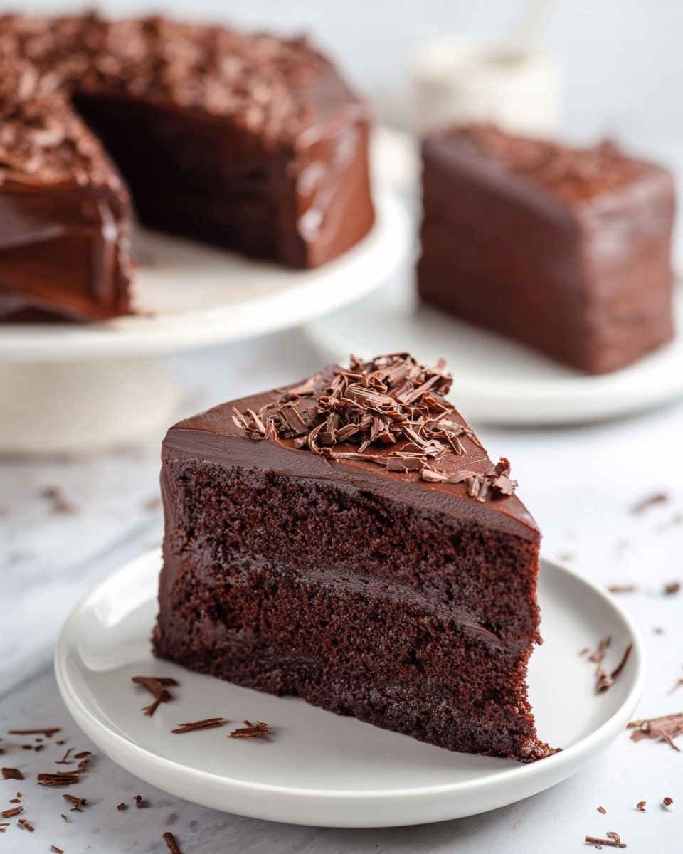 A slice of rich, dark chocolate cake sits on a small white plate, showing three thick, moist layers covered with smooth glossy chocolate frosting. The top layer is decorated with thin, curled chocolate shavings evenly spread. In the background, a matching whole cake with a missing slice and another slice on a white plate are visible, all placed on a white marbled surface with soft natural light highlighting the shiny texture of the frosting. photo taken with an iphone --ar 4:5 --v 7