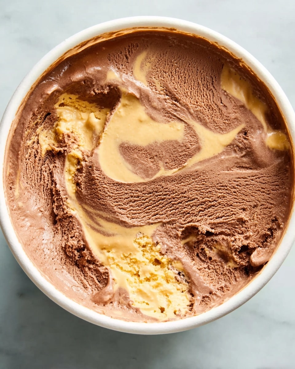 The image shows a white round container with two main layers of ice cream inside. The top layer is a creamy chocolate brown color with a smooth but slightly textured surface. The second layer is a light golden caramel color mixed unevenly with the chocolate, creating a swirled pattern throughout the ice cream. The ice cream has a soft, slightly melted look around the edges. The container sits on a white marbled surface. photo taken with an iphone --ar 4:5 --v 7