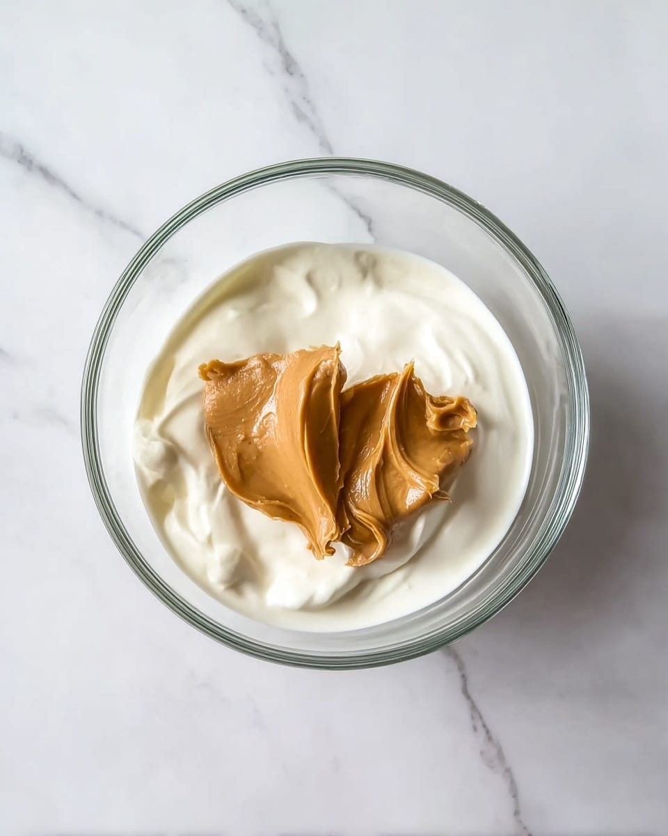 A clear glass bowl sits on a white marbled surface, containing two layers of food. The bottom layer is a smooth, creamy white yogurt or similar white cream, with soft texture and gentle folds. On top, there are two dollops of light brown peanut butter, thick and creamy with visible swirls and ridges. The bowl is photographed from above, showing the contrast between the white cream and the brown peanut butter clearly. photo taken with an iphone --ar 4:5 --v 7