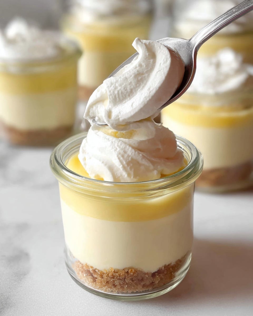 The image shows a small clear glass jar with three visible layers. The bottom layer is a crumbly light brown base, the middle layer is a smooth light yellow custard, and the top layer is a thick, fluffy white whipped cream. A silver spoon is placing a dollop of whipped cream on top of the jar. In the background, there are more jars with similar layers on a white marbled surface. photo taken with an iphone --ar 4:5 --v 7