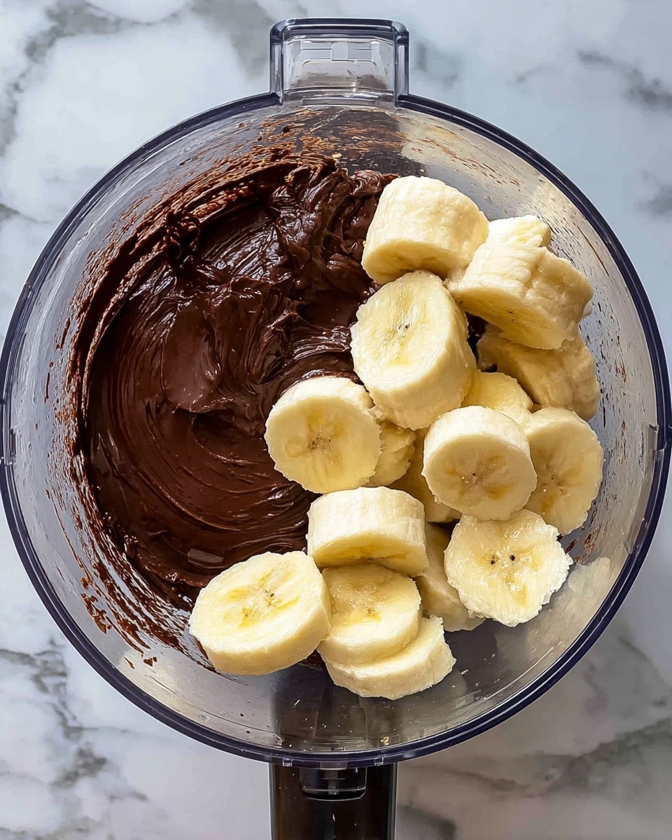 The image shows a top view inside a clear food processor bowl which holds two main layers: on one side, there is a thick, dark brown chocolate spread with a smooth and shiny texture; on the other side, there are several light yellow, sliced banana pieces with soft, slightly textured surfaces. The chocolate is spread over half the base of the bowl, while the banana slices cover the other half. The food processor is placed on a white marbled surface, and the overall look focuses on the contrasting colors and textures inside the bowl. Photo taken with an iphone --ar 4:5 --v 7
