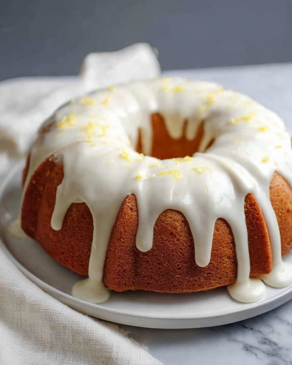 A round bundt cake with a golden brown color sits on a white plate against a white marbled surface. The cake has a thick layer of white icing flowing smoothly over the top and down the sides, with some icing pooling slightly at the bottom edge. There are tiny yellow specks scattered on the icing, adding subtle color. A soft white cloth is draped near the cake's base, creating a cozy feel. Photo taken with an iphone --ar 4:5 --v 7