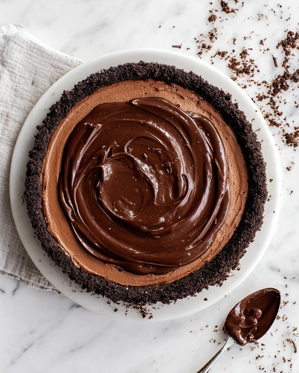 The image shows a triple-layered chocolate tart on a white plate resting on a white marbled surface. The bottom layer is a dark brown crumbly chocolate crust forming a thick edge around the tart. The middle layer is a smooth, lighter brown chocolate mousse filling that fills the crust almost to the top edge. The top layer is a glossy dark chocolate ganache spread evenly with swirling texture on the surface. Around the tart, some chocolate crumbs are scattered. To the right, there is a white spoon with a small amount of the dark chocolate ganache on it. Photo taken with an iphone --ar 4:5 --v 7