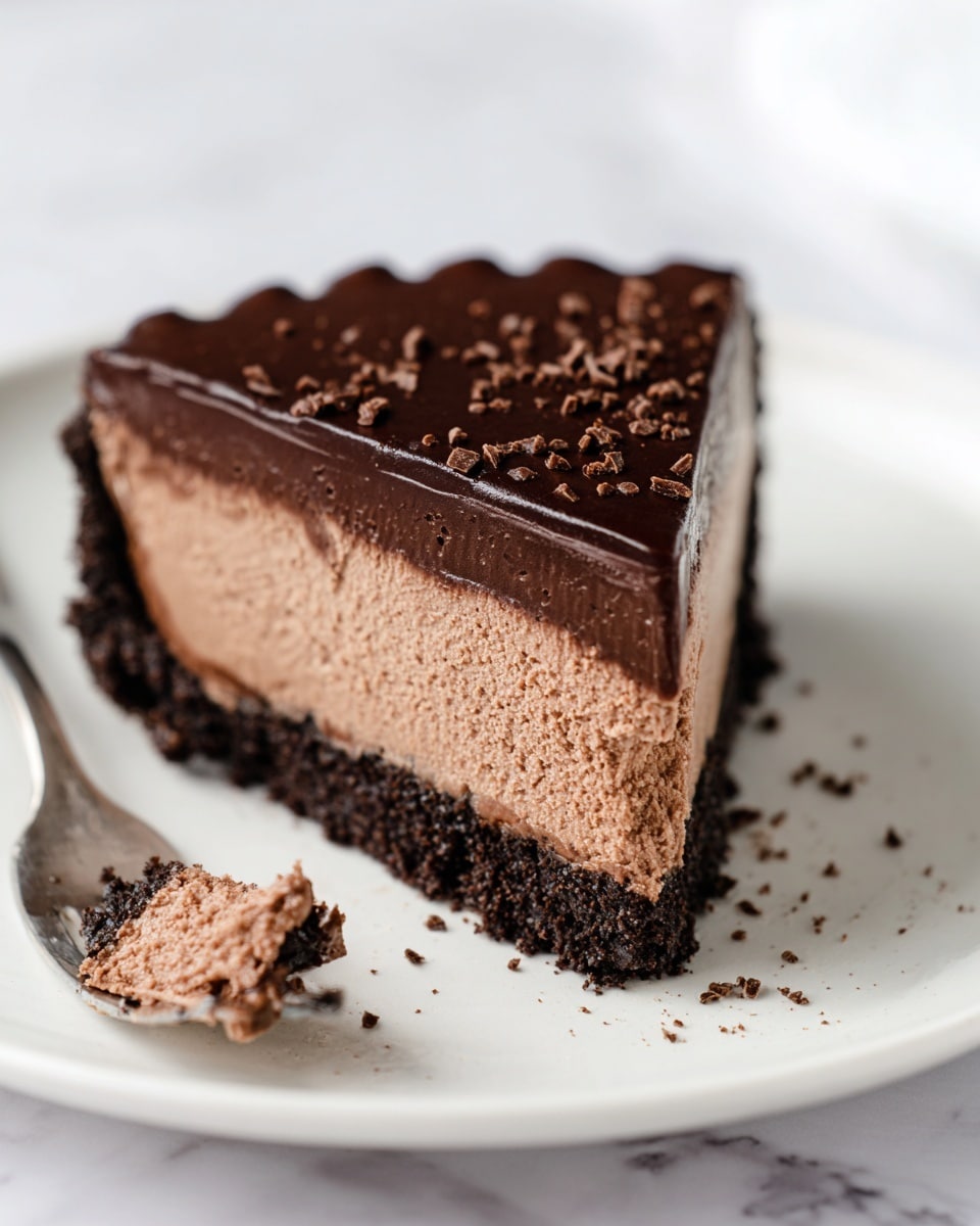 A single slice of chocolate mousse pie sits on a white plate with a few crumbs scattered around. The pie has three layers: a bottom layer of dark chocolate crumb crust, a thick middle layer of light chocolate mousse with a smooth but airy texture, and a shiny, dark chocolate ganache topping. The edges of the crust are slightly ridged, and there are tiny chocolate crumbs sprinkled on top of the ganache. Next to the slice, a fork holds a small piece of the pie, showing the dark crust and light mousse inside. The background is a white marbled texture. photo taken with an iphone --ar 4:5 --v 7