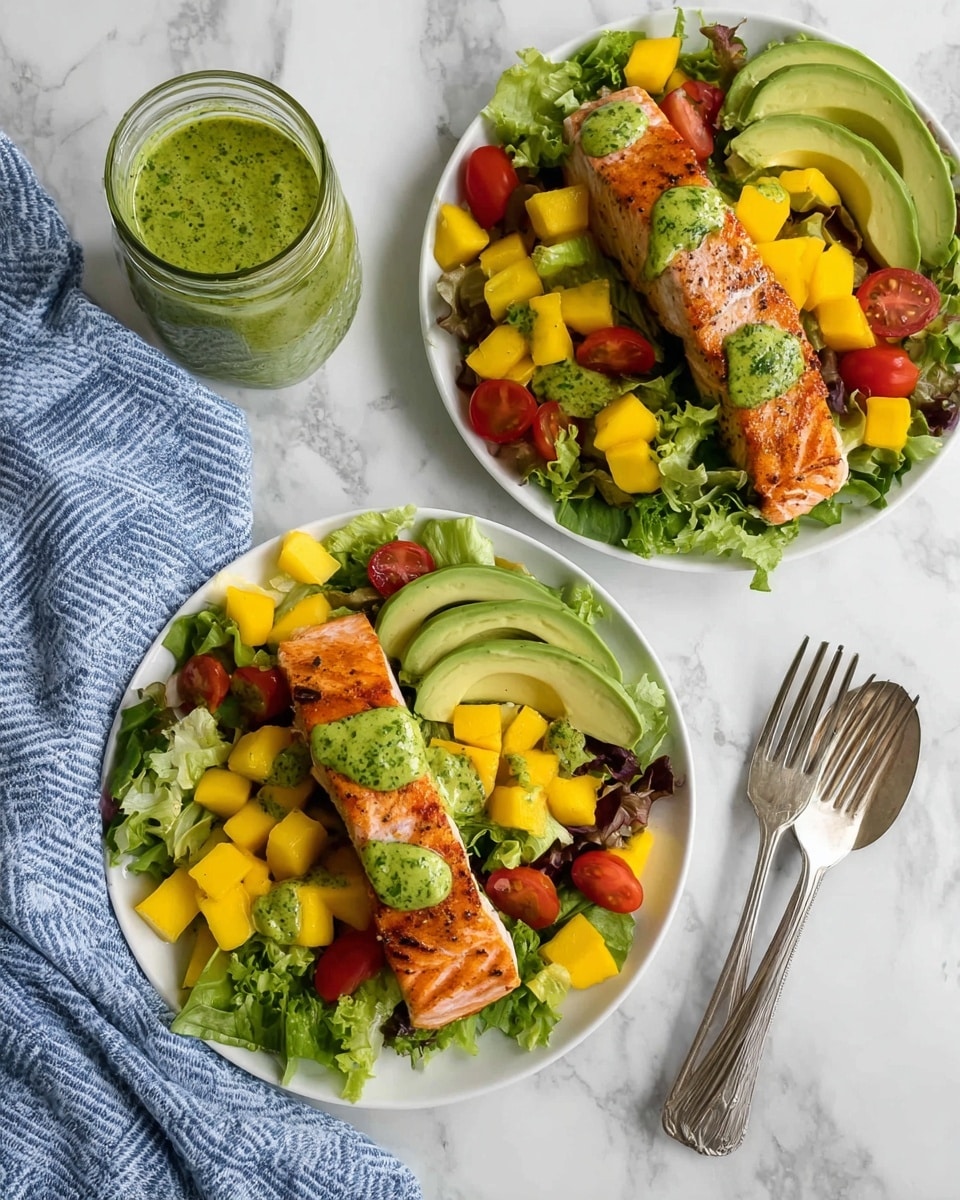 Two white plates sit on a white marbled surface, each with a fresh salad topped with a cooked salmon fillet. Each salad has a green lettuce base with vibrant yellow mango cubes, halved red cherry tomatoes, sliced green avocado fanned out on the side, and is drizzled with a green herb dressing. The salmon fillets are golden-brown with a slight crisp, with green dressing drops on top. A jar of the same green dressing with a spoon inside is placed beside the plates. Two forks rest next to a blue and white striped cloth napkin. photo taken with an iphone --ar 4:5 --v 7