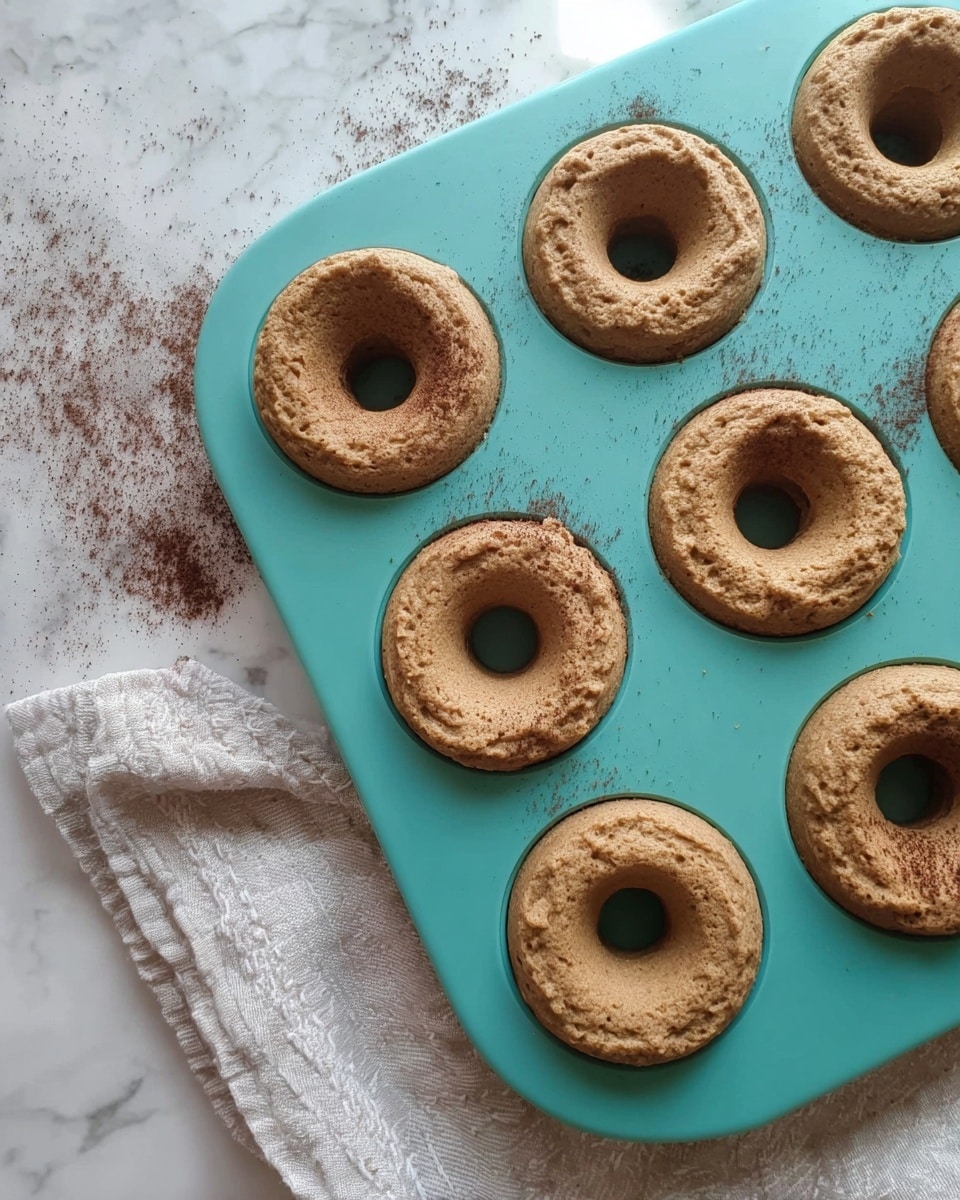 A teal silicone donut baking tray holds nine freshly baked light brown donuts with a slightly cracked and rough texture. Each donut has a small round hole in the center, and some have a little excess dough around the edges. Cocoa powder is lightly dusted on a few donuts and the tray. A soft white cloth with a faint pattern is partially visible underneath the tray on one side. The scene is set on a white marbled surface photo taken with an iphone --ar 4:5 --v 7