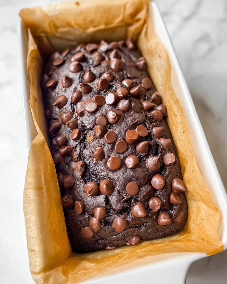 A single thick dark brown chocolate cake loaf is shown inside a white ceramic baking dish lined with light brown parchment paper. The top layer is covered with many shiny medium-sized milk chocolate chips that are scattered unevenly, creating a rich and textured look. The cake surface beneath the chocolate chips is slightly cracked, showing a moist and dense texture inside. The edges of the parchment paper rise up and over the sides of the baking dish. The photo is taken on a white marbled surface photo taken with an iphone --ar 4:5 --v 7