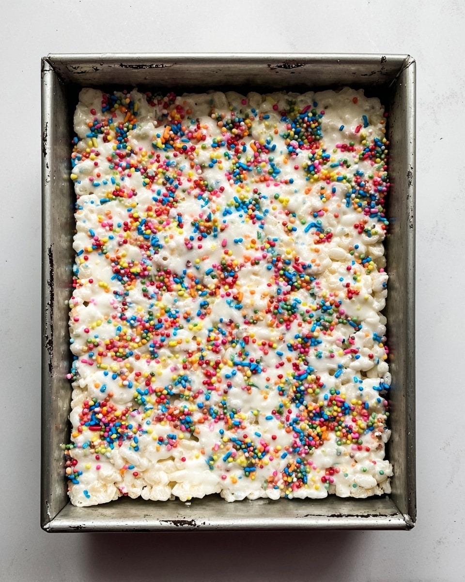 The image shows a close-up top view of a single-layer white marshmallow rice treat in a square metal baking pan. The treat is thick, sticky, and covered evenly with colorful round sprinkles scattered all over the top. The pan edges show some slight smudges of the treat mixture. The whole scene is set against a white marbled textured surface. photo taken with an iphone --ar 4:5 --v 7