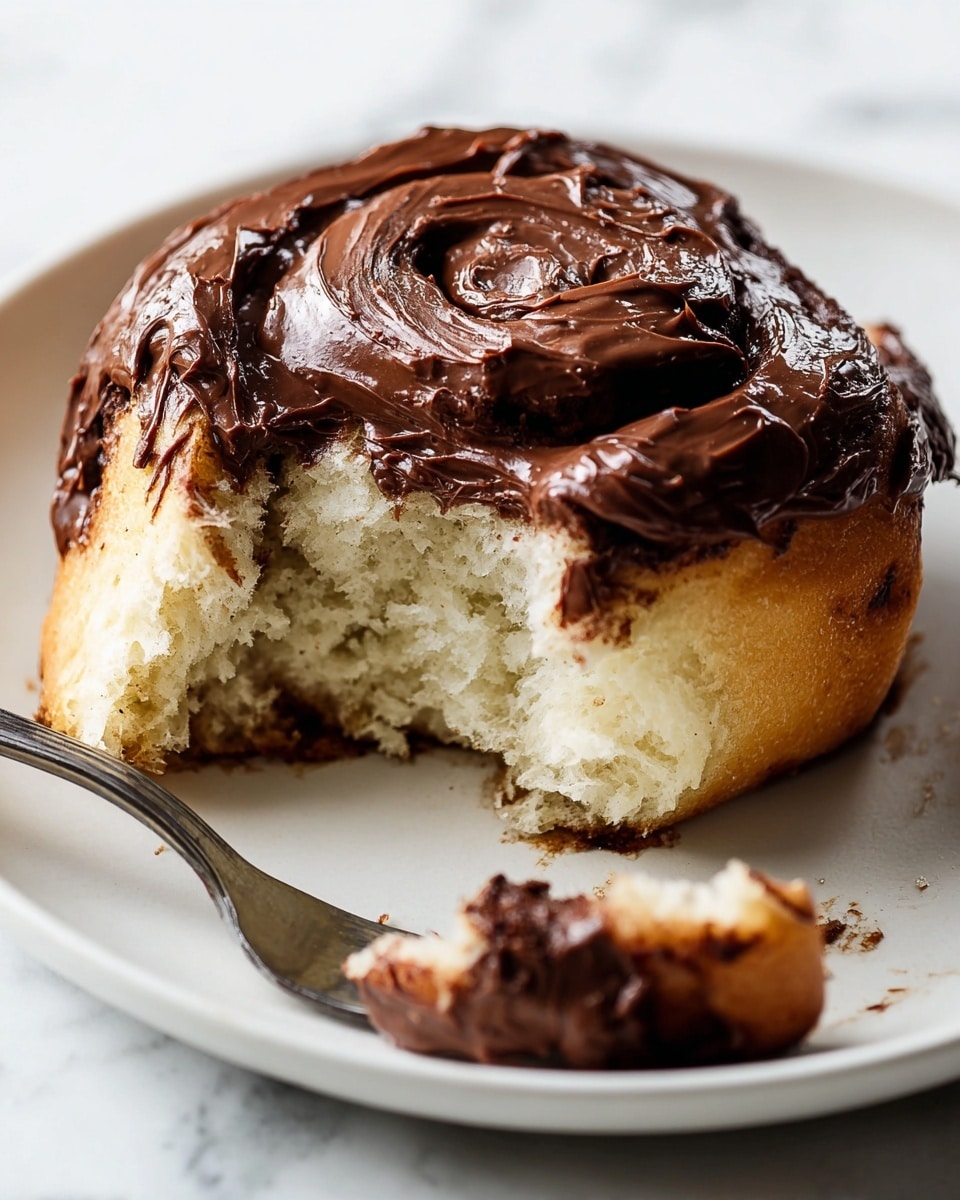 A close-up image shows a single cinnamon roll on a white plate with a glossy, thick layer of dark chocolate spread on top. The roll has a light golden-brown outer layer with soft, fluffy, pale cream inside visible where a piece has been torn off from the front left. The chocolate topping is shiny with smooth swirls, covering the upper spiral of the roll. Next to the cinnamon roll on the plate, a small piece of the roll rests on a silver fork, showing its airy texture and a slightly darker, caramelized edge. The background is a white marbled surface, and the lighting highlights the richness of the chocolate and softness of the dough. photo taken with an iphone --ar 4:5 --v 7