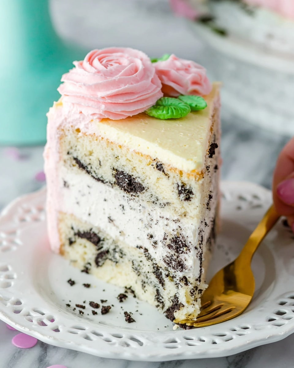 A slice of cookies and cream cake sits on a white plate with a delicate cut-out rim, placed on a white marbled surface. The cake has two thick layers of creamy white cake with dark cookie pieces mixed throughout, separated by a thin layer of white frosting. The outside edge of the cake is coated in light pink frosting, while the top layer has a smooth pale yellow frosting. A large, pink frosting rose decoration with green leaves is on top near the back edge. A gold fork rests at the front of the plate, slightly angled, and a woman's hand is about to pick up the slice. Photo taken with an iphone --ar 4:5 --v 7