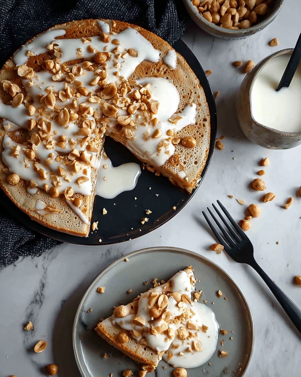 A round light brown pancake with small bubbles on its surface is topped with crunchy golden peanuts scattered all over, drizzled with thick white cream in thin streaks. One slice is taken out from the pancake and placed on a white plate below, showing the same peanut and cream topping on it, with cream pooling slightly beside the slice. The white plate sits on a white marbled surface with extra peanuts scattered around. A black fork and a small cup of white cream with a black spoon are nearby. Photo taken with an iphone --ar 4:5 --v 7