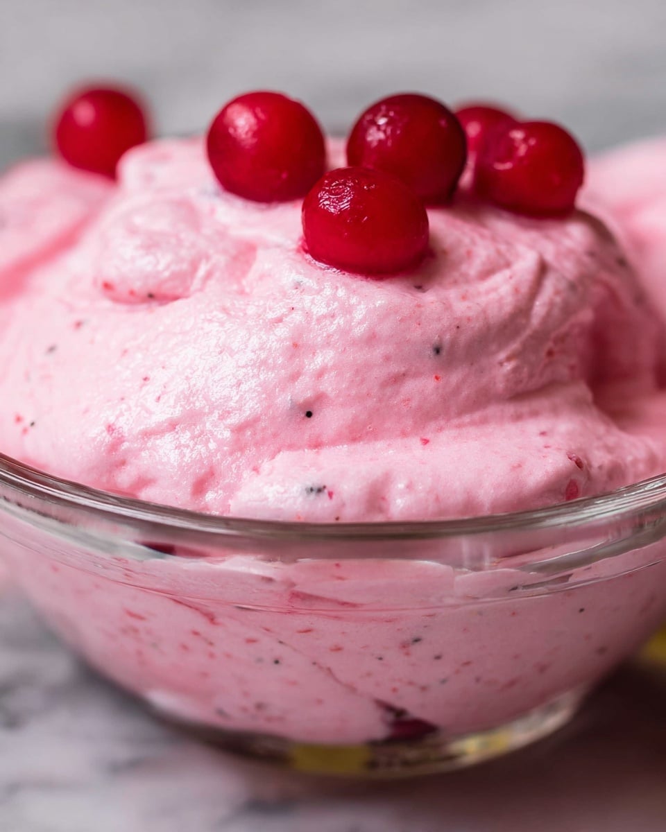 This close-up image shows a smooth, fluffy pink mousse-like dessert with a light and airy texture. The dessert is in a clear glass bowl, and it has small specks of darker pink mixed inside. On top, several small, shiny red berries are placed, sitting gently on the mousse surface. The background is a white marbled texture. photo taken with an iphone --ar 4:5 --v 7