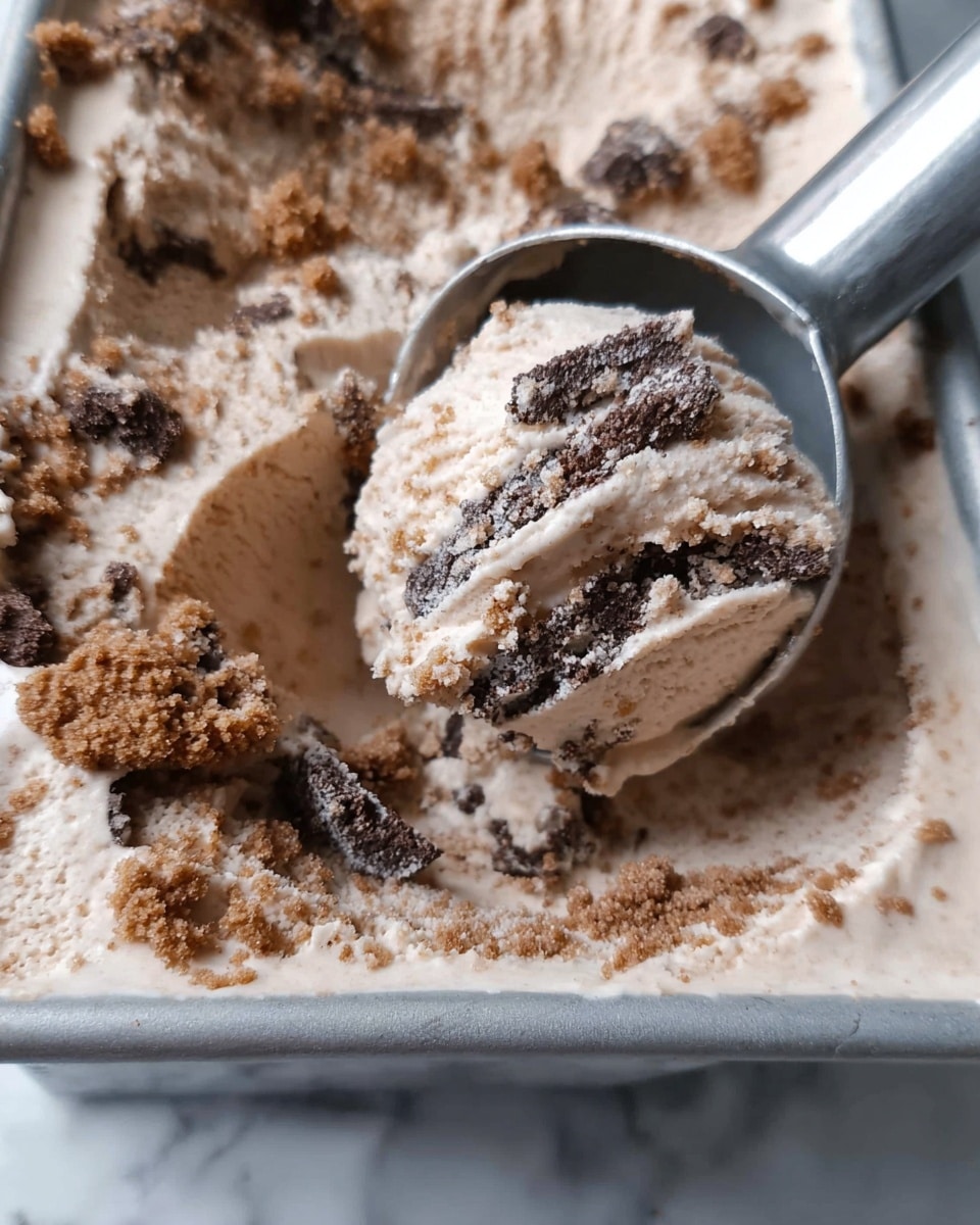The image shows a close-up view of a scoop of light brown ice cream with visible darker brown cookie chunks mixed inside. The ice cream has a creamy texture with some icy crystals, and the darker cookie pieces look crumbly and unevenly spread throughout the scoop. A silver ice cream scoop is in the act of lifting a portion from the container. The container beneath is filled with the same light brown ice cream and cookie piece mixture. The background is a white marbled texture. photo taken with an iphone --ar 4:5 --v 7