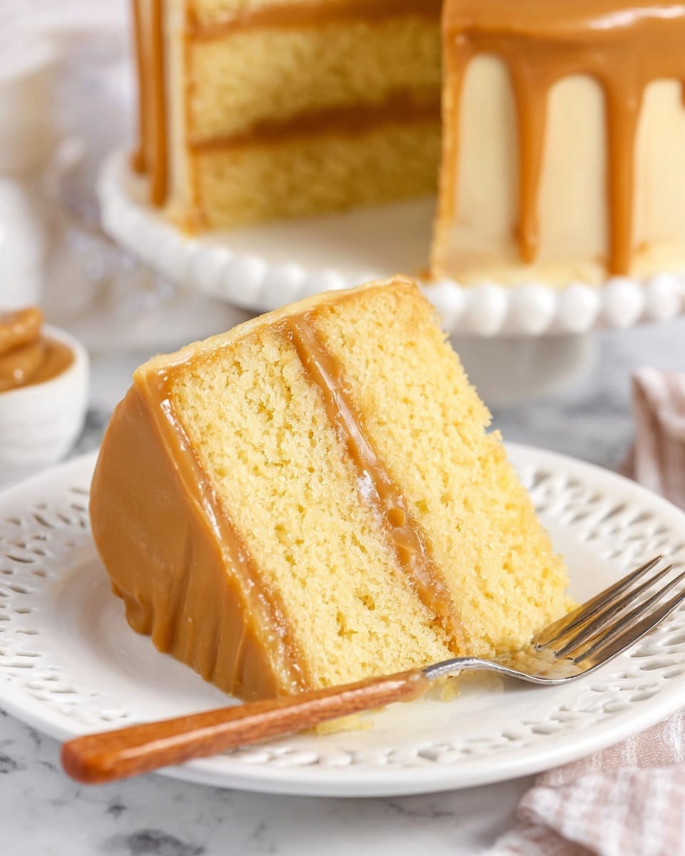 A slice of three-layer yellow cake with smooth light brown caramel frosting between each layer and covering the outside sits on a white plate with a delicate cut-out pattern along the edge. The cake layers look soft and moist with a fine crumb texture, while the caramel frosting is shiny and creamy. There is a fork with a wooden handle next to the slice, and the background shows the rest of the cake on a white marbled surface. Photo taken with an iphone --ar 4:5 --v 7
