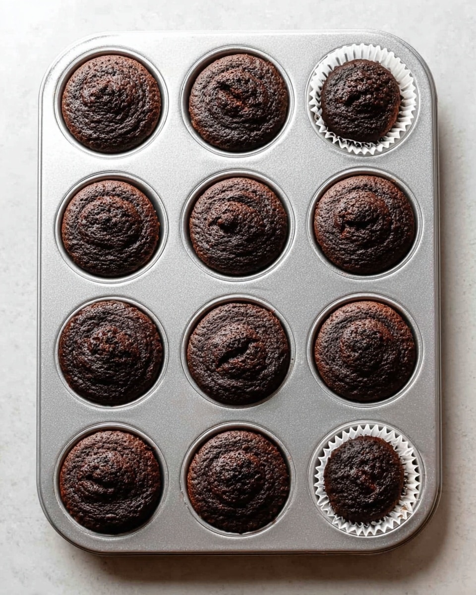 A silver-colored metal muffin tray holds twelve dark chocolate muffins arranged in three rows and four columns. Each muffin is round with a domed, slightly cracked top showing a rich, soft texture. One muffin in the top row, far right, is in a white paper liner, contrasting with the others that are directly in the tray cups. The tray sits on a white marbled surface, with light shining softly to bring out the deep brown color of the muffins. photo taken with an iphone --ar 4:5 --v 7