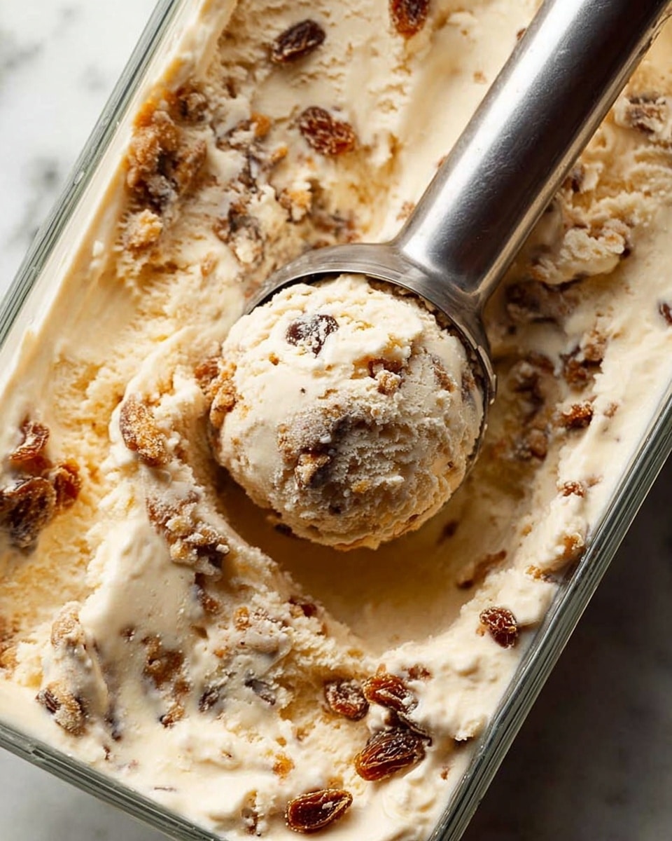 The image shows a close-up of creamy beige ice cream mixed with small chunks of brown raisins and cake pieces spread evenly in a clear rectangular glass container. A metal ice cream scoop is partially submerged, lifting a round, textured scoop full of the same creamy ice cream and mix-ins. The ice cream looks smooth with rough, chunky bits giving it a mixed texture. The background surface is a white marbled texture. photo taken with an iphone --ar 4:5 --v 7