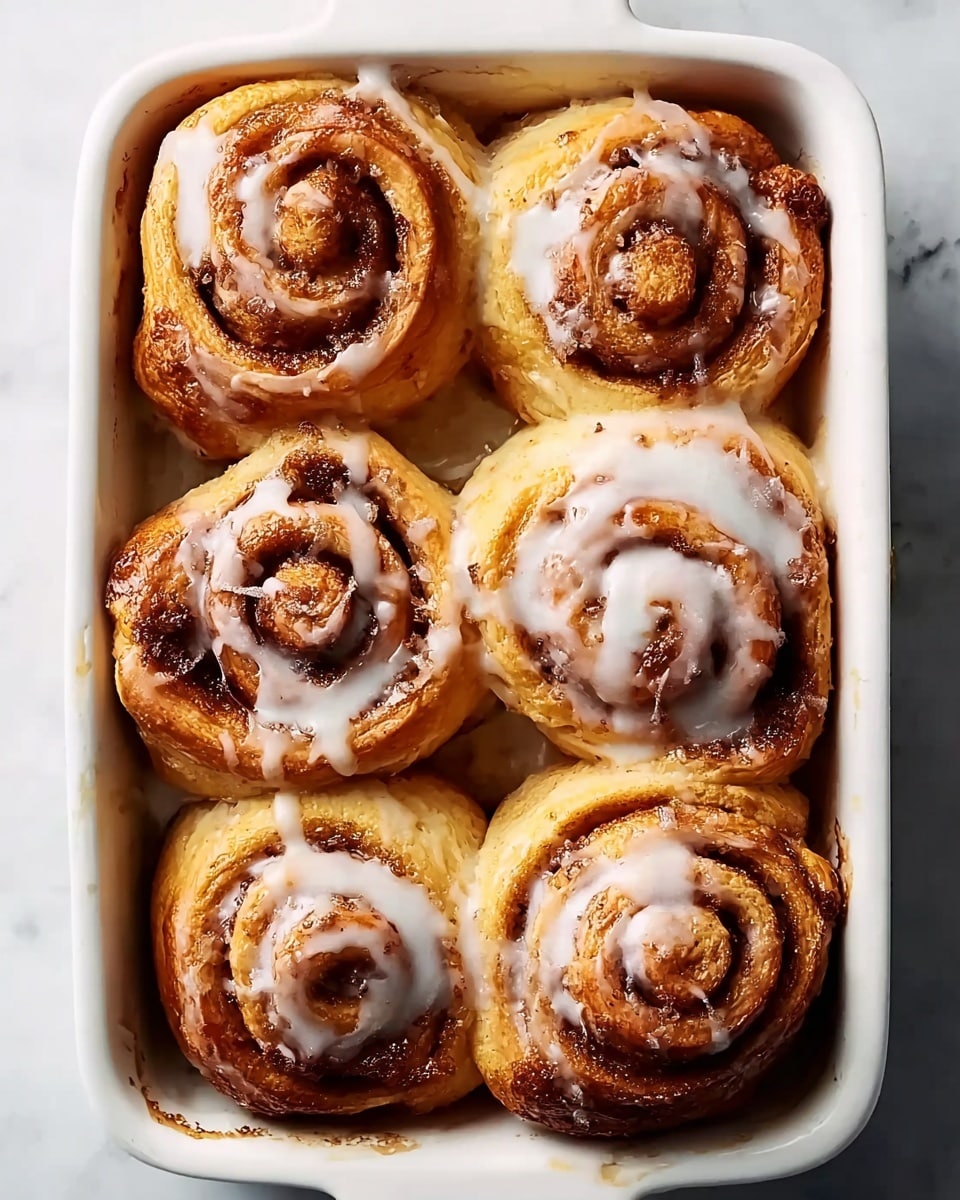 Six cinnamon rolls sit closely in a white rectangular baking dish on a white marbled surface. Each roll is golden brown with a visible swirl of darker cinnamon filling spiraling from the center outwards. A light white icing drizzle covers the tops unevenly, adding a glossy texture that contrasts with the soft dough. The rolls look warm and fluffy, slightly touching each other, filling the dish almost completely. photo taken with an iphone --ar 4:5 --v 7