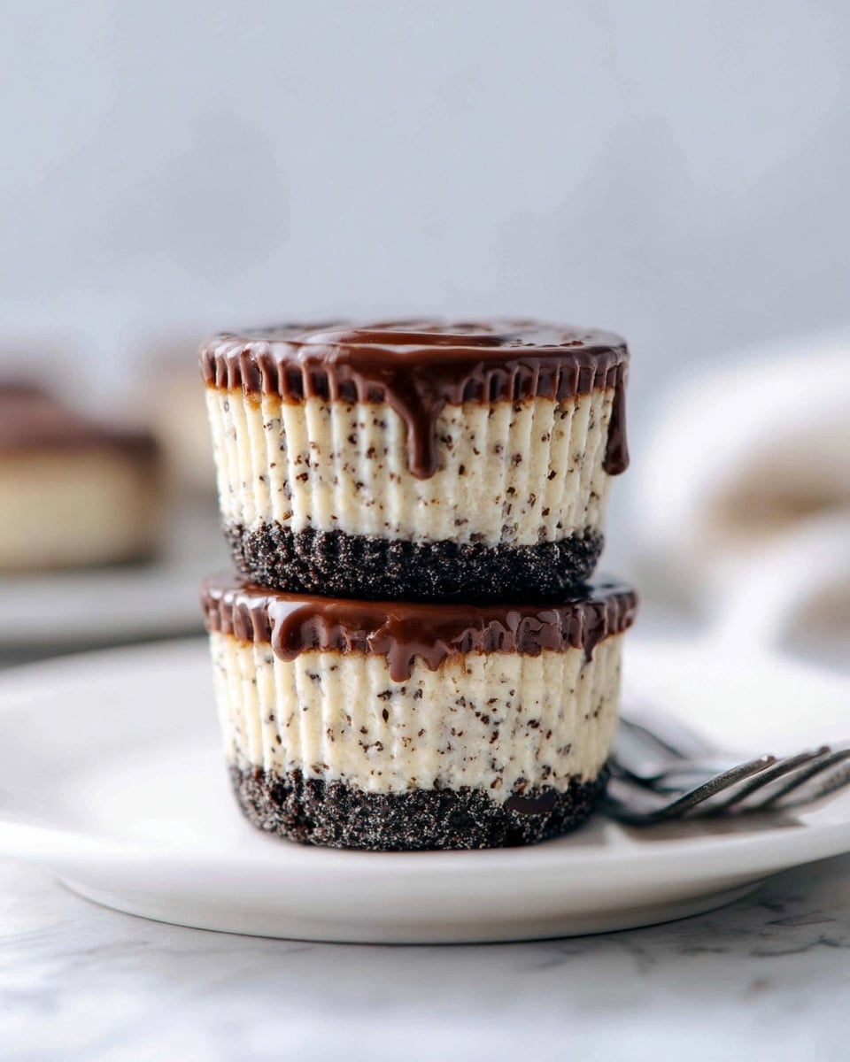 The image shows two small cakes stacked on a white plate. Each cake has three layers: a dark, crumbly base at the bottom, a thick middle layer that is light cream color with small dark specks all over, and a smooth, shiny chocolate layer on top that slightly drips down the sides. The cakes are in white paper cups with visible ridges. The background and surface are white marbled texture, and there is a blurred fork next to the plate. Photo taken with an iphone --ar 4:5 --v 7