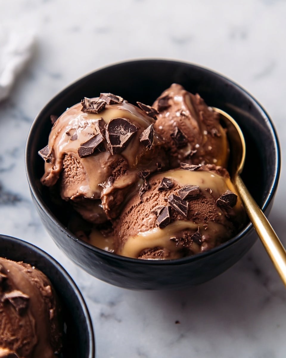 The image shows a black bowl filled with three scoops of chocolate ice cream. The ice cream is rich brown, smooth, and creamy with a slightly melting texture. It is topped with a light brown sauce drizzled unevenly across the scoops, and small pieces of dark chocolate are sprinkled on top, adding texture. A shiny gold spoon rests inside the bowl, touching the ice cream, and the bowl is placed on a white marbled surface. Another bowl with similar ice cream is partly visible at the bottom left corner. photo taken with an iphone --ar 4:5 --v 7