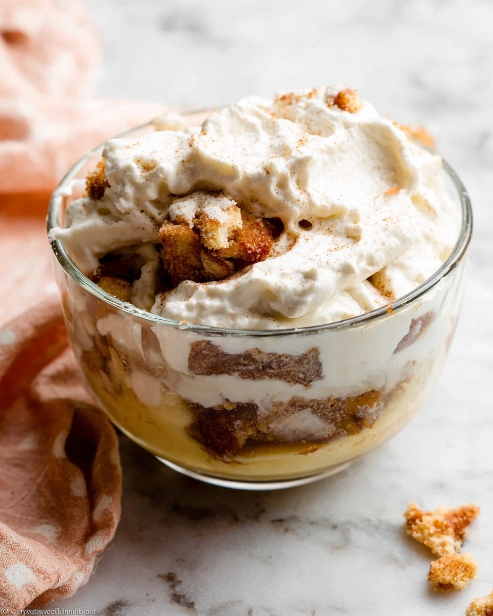 The image shows a clear glass bowl filled with a layered dessert. At the bottom, there is a crumbly brown layer with some chunks that look like cake or cookie pieces. Above that is a thick yellowish creamy layer with a smooth texture. The top layer is a fluffy white cream with some visible specks, likely cinnamon or vanilla. The layers are mixed together slightly, creating a soft, rich appearance. The bowl sits on a white marbled surface with a light peach and white cloth nearby. Photo taken with an iphone --ar 4:5 --v 7