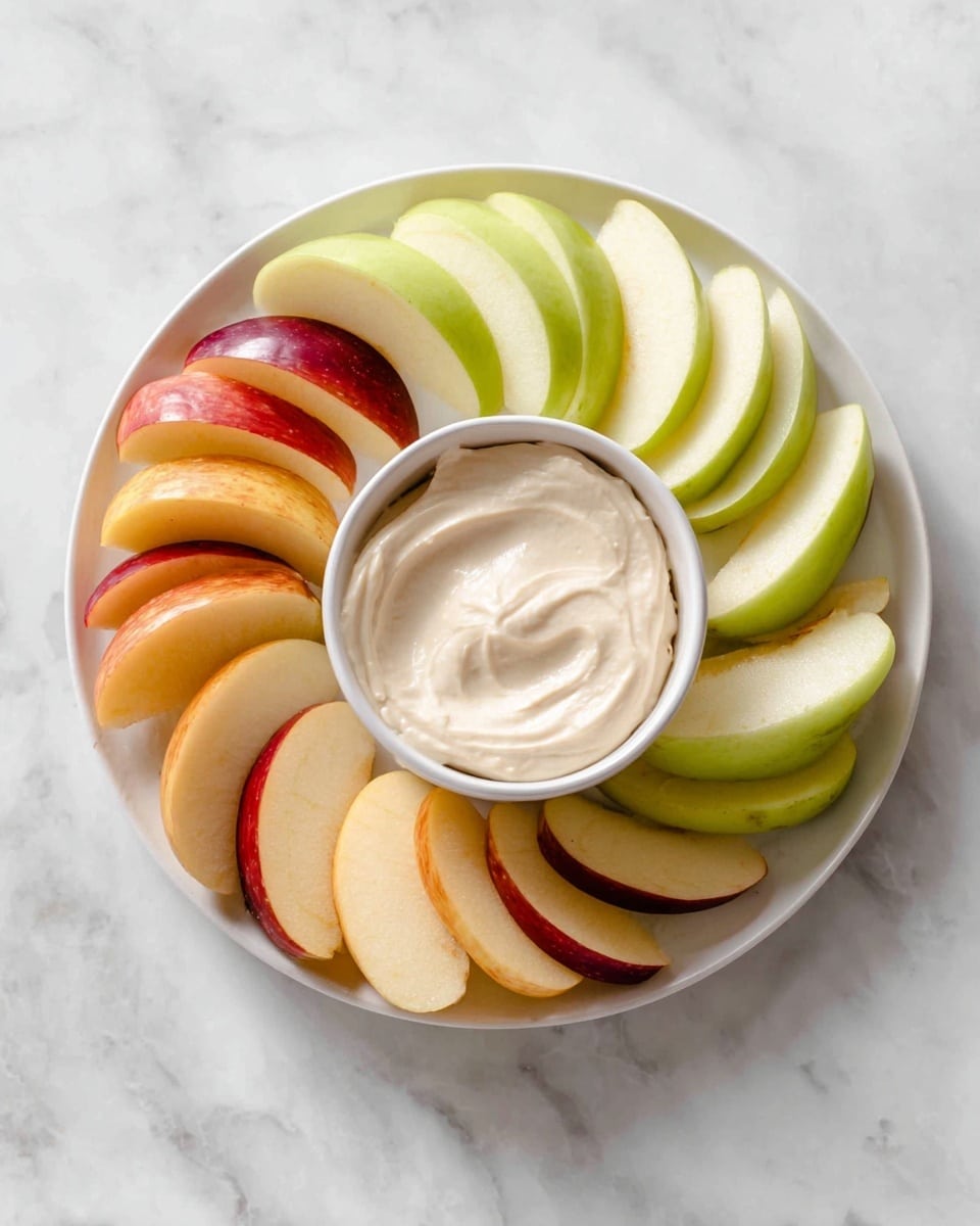 A white plate on a white marbled surface holds thin, evenly sliced apple pieces arranged in three groups around a small white bowl of creamy off-white dip. One group of apple slices has a bright green skin, another has a yellowish-red skin, and the last has a red skin with a hint of yellow. The smooth dip forms a gentle swirl inside the bowl and is centered on the plate. The apple slices fan outwards, slightly overlapping each other, making a colorful and fresh presentation photo taken with an iphone --ar 4:5 --v 7