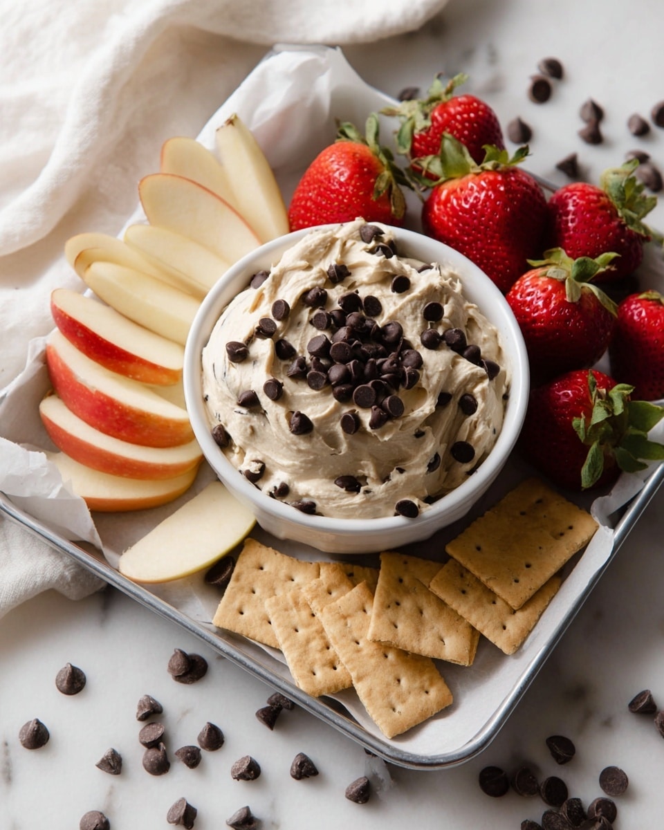 A white small bowl in the center holds a thick, creamy dip with a light beige color, topped with many small dark chocolate chips. Surrounding the bowl on a white metal tray lined with white paper are three groups of snacks: bright red and green strawberries on the top right, fresh apple slices with red and yellow skin on both sides, and several light brown rectangular crackers at the bottom. Some chocolate chips are scattered around the tray on a white marbled surface. A white cloth is partially visible on the left side. Photo taken with an iphone --ar 4:5 --v 7