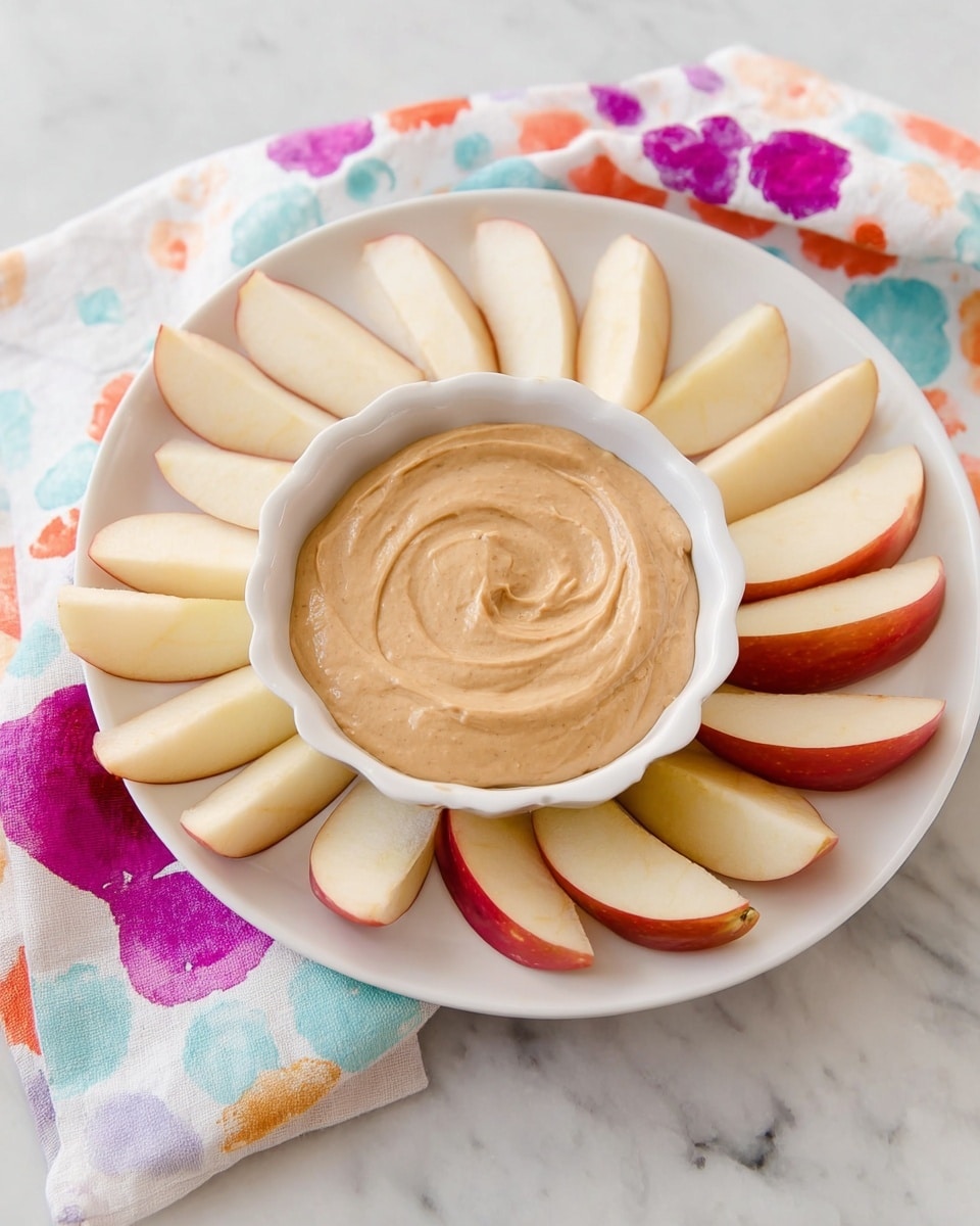 A white plate on a white marbled surface holds thin apple slices arranged in a circular pattern from left to right. In the center of the plate is a white scalloped bowl filled with smooth, light brown creamy dip that has gentle swirls on its surface. To the left side of the plate is a cloth with a white base and colorful abstract patterns, adding a soft touch to the setting. The overall look is fresh, simple, and inviting. photo taken with an iphone --ar 4:5 --v 7