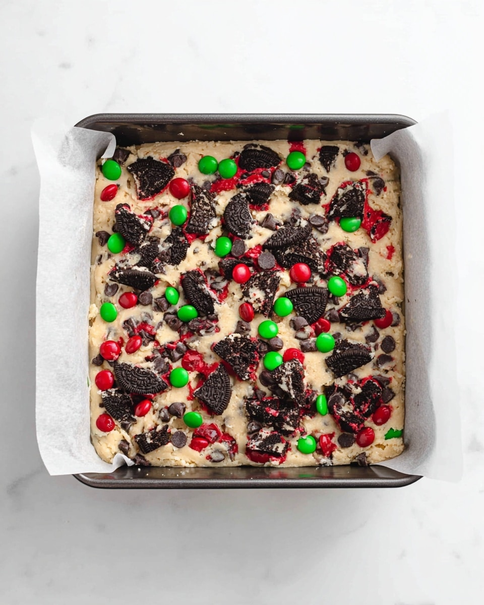 A square baking pan lined with white parchment paper holds a thick cookie dough layer that is pale beige with a slightly rough texture. The dough is generously topped with broken black sandwich cookie pieces that have bright red filling showing in some spots, along with scattered red and green candy-coated chocolates, and many small dark chocolate chips sprinkled evenly across the top. The pan rests on a white marbled surface. photo taken with an iphone --ar 4:5 --v 7
