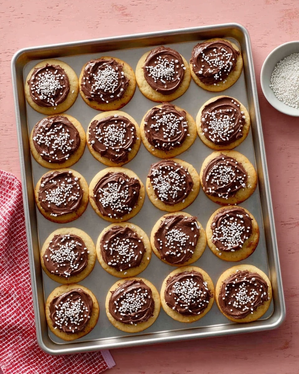 A metal baking tray holds 20 round cookies arranged in 4 rows and 5 columns, each cookie having a light golden base topped with a smooth layer of dark brown chocolate frosting. The frosting is spread unevenly, creating a textured surface, and each cookie is decorated with white sprinkles in different shapes, including small rods and dots. The tray rests on a pink surface with a red-and-white checkered cloth partially visible on the left side. In the upper right corner of the image, a small white bowl contains more white sprinkles. Photo taken with an iphone --ar 4:5 --v 7