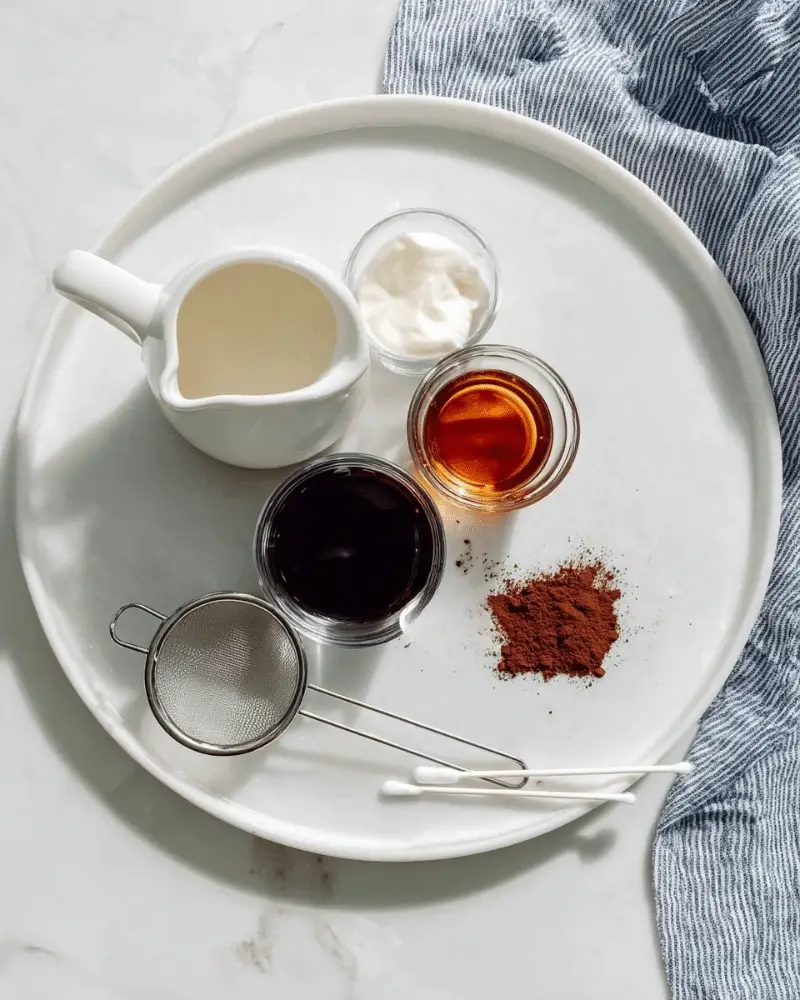 A white round plate on a white marbled surface holds four small containers with different liquids: one white ceramic pitcher with off-white half and half, a clear glass with dark brown Kahlua, a clear glass with amber-colored amaretto, and a small silver metal cup with clear vanilla vodka. Nearby, a metal sieve contains some loose cocoa powder. Two white-tipped cotton swabs lie on the plate near the bottom right, and a blue and white striped cloth is partially visible at the bottom right corner. Photo taken with an iphone --ar 4:5 --v 7