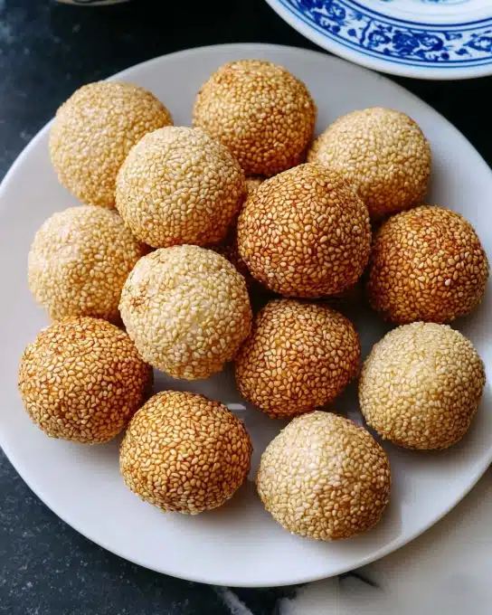 A white plate holds about fifteen small, round balls that are evenly coated with golden-brown sesame seeds, giving them a textured, crunchy look. Each ball is smooth beneath the sesame layer and varies slightly in color from pale beige to a light golden brown, showing they are baked or toasted. The balls are arranged close together in the center of the plate, which rests on a dark surface with a white marbled texture seen at the edge. Part of a white bowl with blue patterns is visible in the top right corner. photo taken with an iphone --ar 4:5 --v 7
