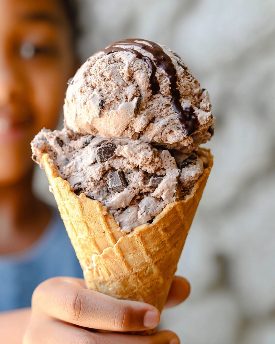 A close-up of a large scoop of chocolate ice cream with visible chunks mixed in, resting on a waffle cone that has a drizzle of dark chocolate near its top edge. The ice cream appears creamy with a slightly rough texture showing the chunks inside. A woman's hand is holding the waffle cone from the bottom, and there is a soft blurred background with a hint of a person's face. The whole scene is set against a white marbled texture background. photo taken with an iphone --ar 4:5 --v 7