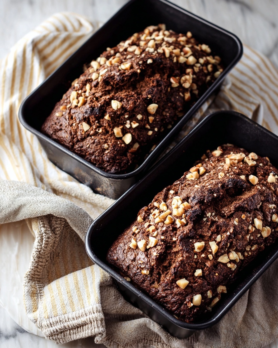 Two dark brown loaves of bread with rough cracked tops covered in light brown chopped nuts sit inside black baking pans. The texture on the bread tops looks crunchy and crumbly. The pans rest on a cloth with pale yellow, white, and grey stripes, which is laid on a white marbled surface. A pale beige oven mitt is placed near the front. The photo is taken with an iphone --ar 4:5 --v 7