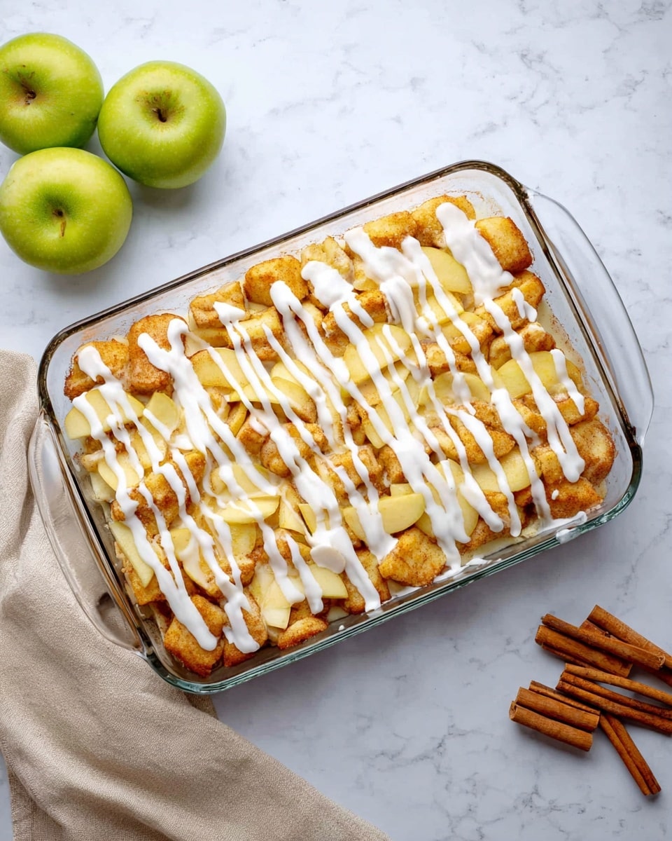 The image shows a clear glass rectangular baking dish filled with a layered dessert. The bottom layer is made of small, golden brown dough pieces that are fluffy and slightly uneven in shape. On top of this is a layer of pale yellow apple slices, spread across evenly. The whole dish is drizzled with white icing in thick vertical lines covering the top fully. The dish sits on a white marbled surface, with a light beige cloth folded nearby holding three green apples and three cinnamon sticks on the right side of the image. Photo taken with an iphone --ar 4:5 --v 7