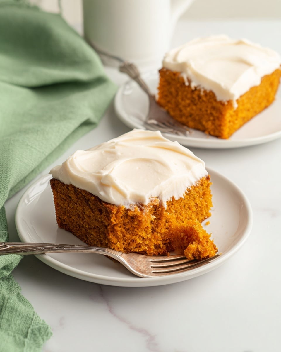 The image shows two pieces of orange-colored cake with a thick, smooth layer of white frosting on top. Each piece is on a small white plate, with a silver fork resting on the plate. One piece in the front has a small bite taken out, showing the soft, moist texture of the cake inside. The background is a clean white marbled surface, and a soft green cloth is partially visible in the top left corner. The photo taken with an iphone --ar 4:5 --v 7