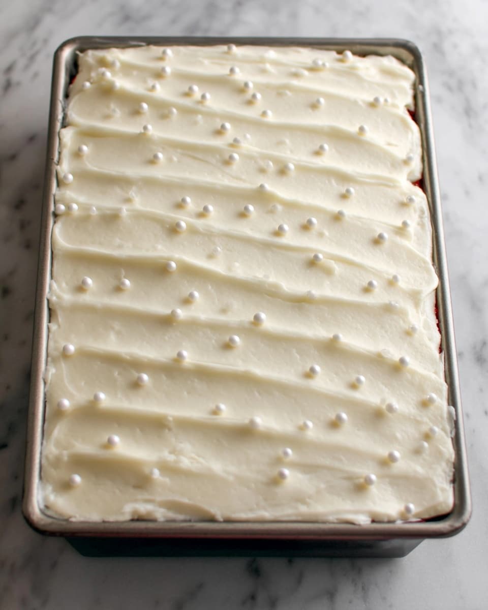 The image shows a rectangular cake with one visible layer in a metal pan. The cake is covered with a thick layer of smooth, creamy white frosting spread evenly on top and around the edges, with subtle lines creating a gentle texture. Small, round white edible pearls are scattered across the frosting, adding slight dimension and decoration. The cake sits on a white marbled surface. photo taken with an iphone --ar 4:5 --v 7
