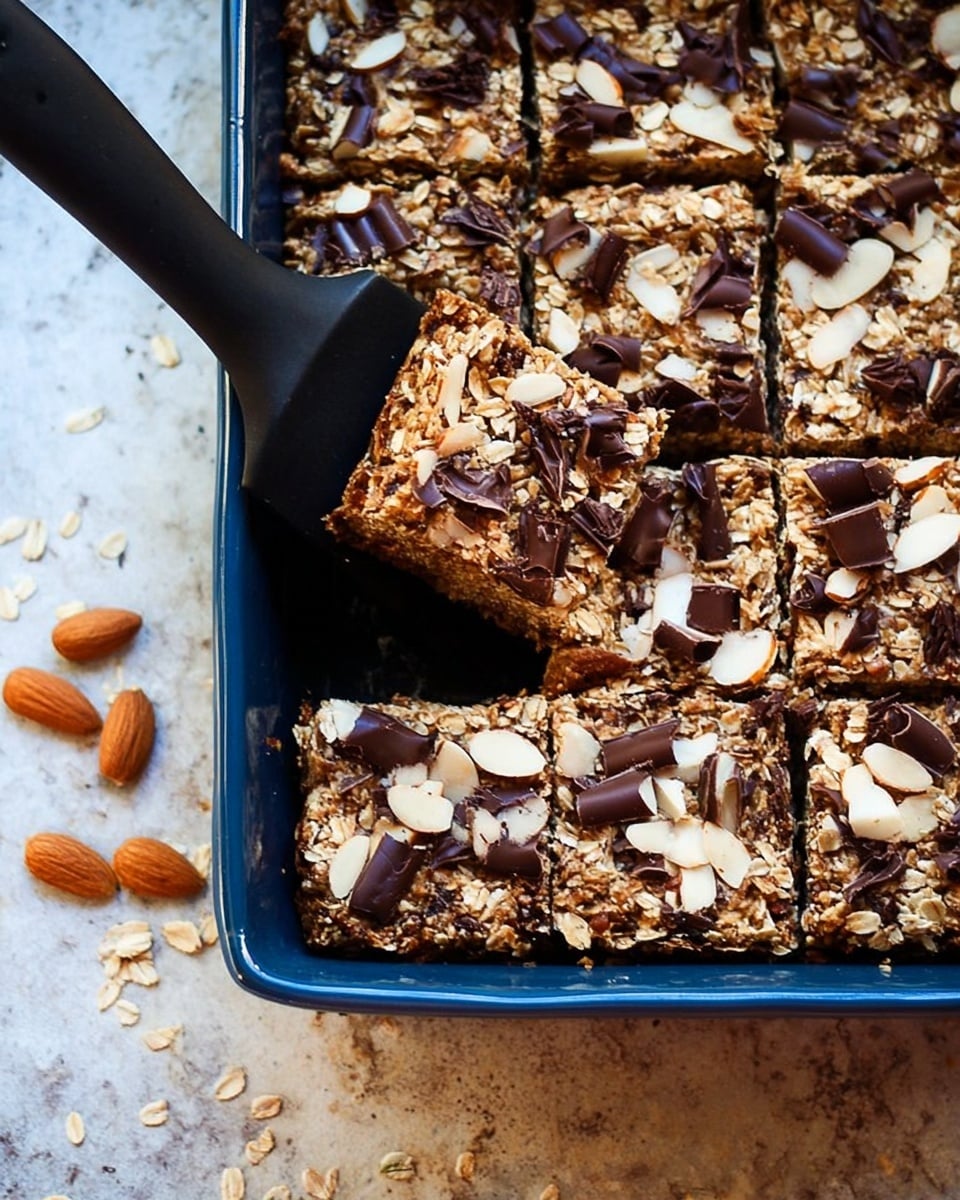 The image shows a dark blue baking dish filled with rectangular oat bars cut into squares. The bars have a textured, light brown oat base layer with scattered sliced almonds and curled dark chocolate pieces on top. One bar is being lifted out with a black spatula, exposing the inside of the oat bar which matches the top's light brown color and oats. The baking dish is placed on a white marbled textured surface, with some almond slices and oats scattered around. Photo taken with an iphone --ar 4:5 --v 7