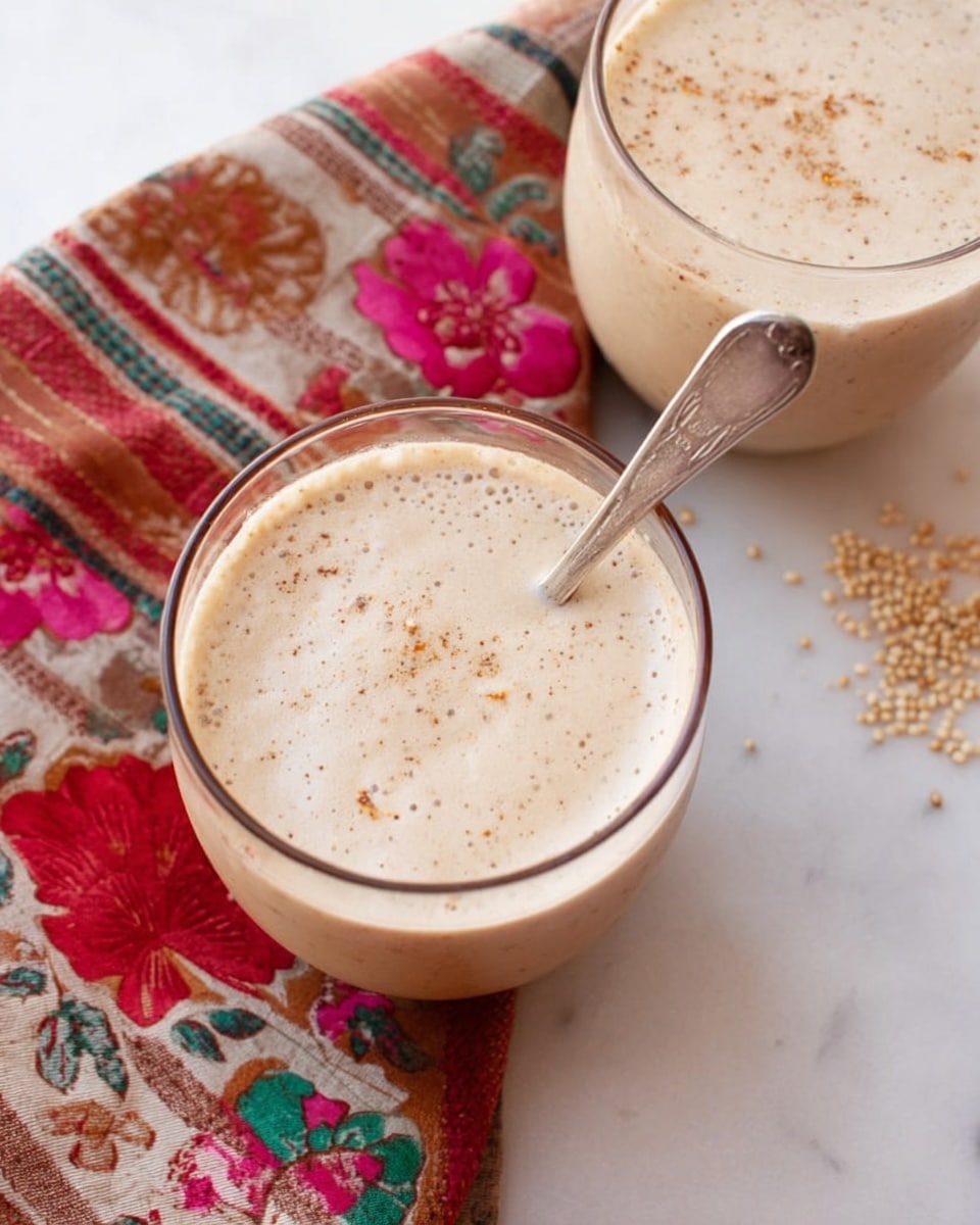 Two clear round glasses filled with a smooth, creamy beige drink that has tiny bubbles and light brown specks swirled on top, one glass holding a silver spoon inside. The glasses rest on a white marbled surface next to a colorful cloth with floral and striped patterns in red, pink, green, and brown. Small beige grains are scattered on the surface near the glasses. Photo taken with an iphone --ar 4:5 --v 7