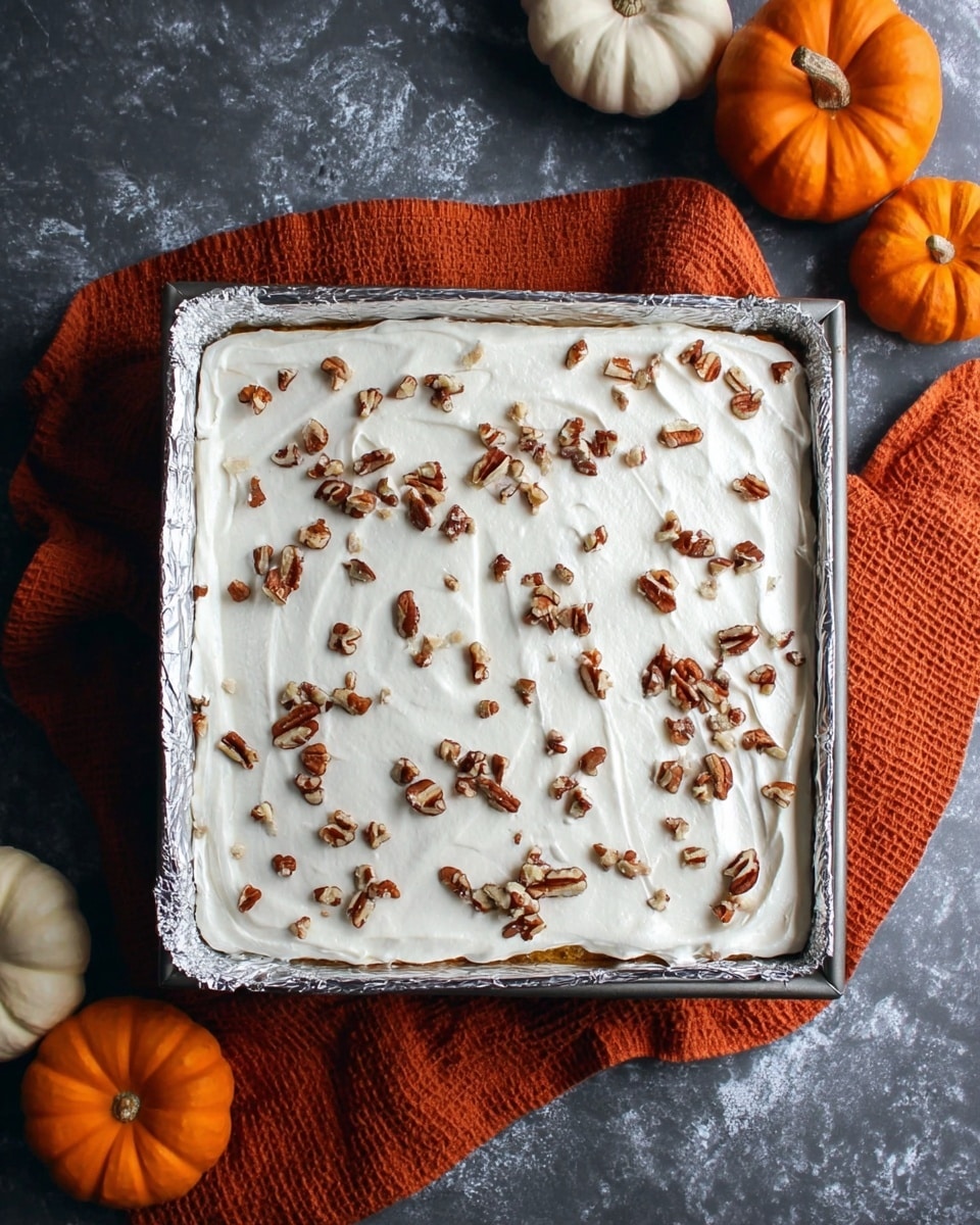 A square baking pan lined with silver foil holds a dessert with two visible layers: the bottom layer is a light brown cake or crust, and the top layer is a thick, white creamy frosting spread evenly across the surface. Small pieces of toasted pecans are scattered on top, adding texture and a darker brown color contrast. The pan is placed on a dark gray surface with a white marbled texture, and an orange textured cloth is partly visible under the pan. Around the pan, there are orange and white pumpkins adding a fall or autumn theme. photo taken with an iphone --ar 4:5 --v 7
