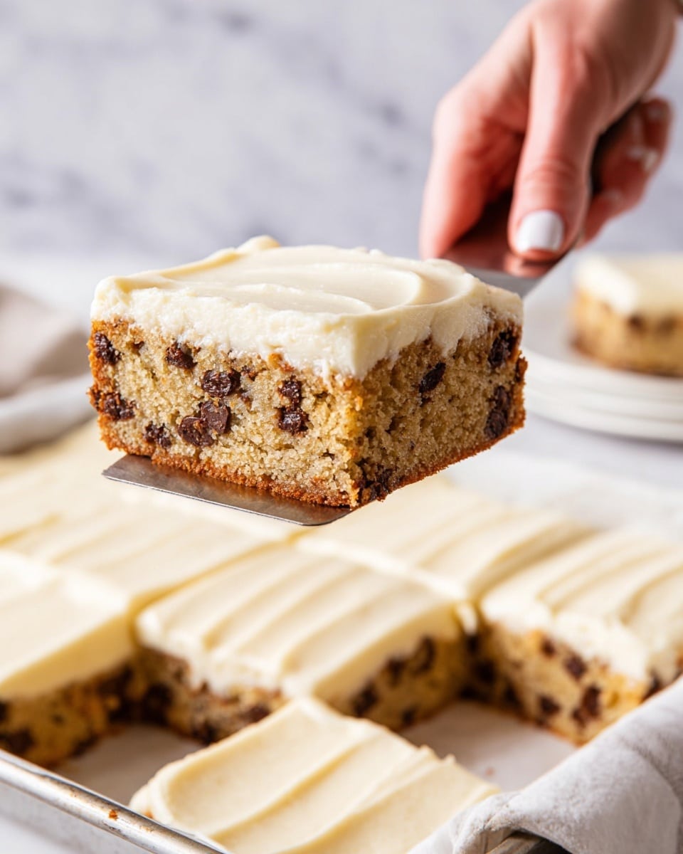 A square slice of chocolate chip cake with a thick top layer of smooth, creamy white frosting is being lifted with a spatula by a woman's hand. The cake layer is light brown with many small dark chocolate chips spread evenly throughout. The slice is thick and looks soft, resting above a white baking tray filled with more frosted cake squares. The background has a white marbled texture, and there is a plain white plate with another frosted cake square blurred out behind. Photo taken with an iphone --ar 4:5 --v 7