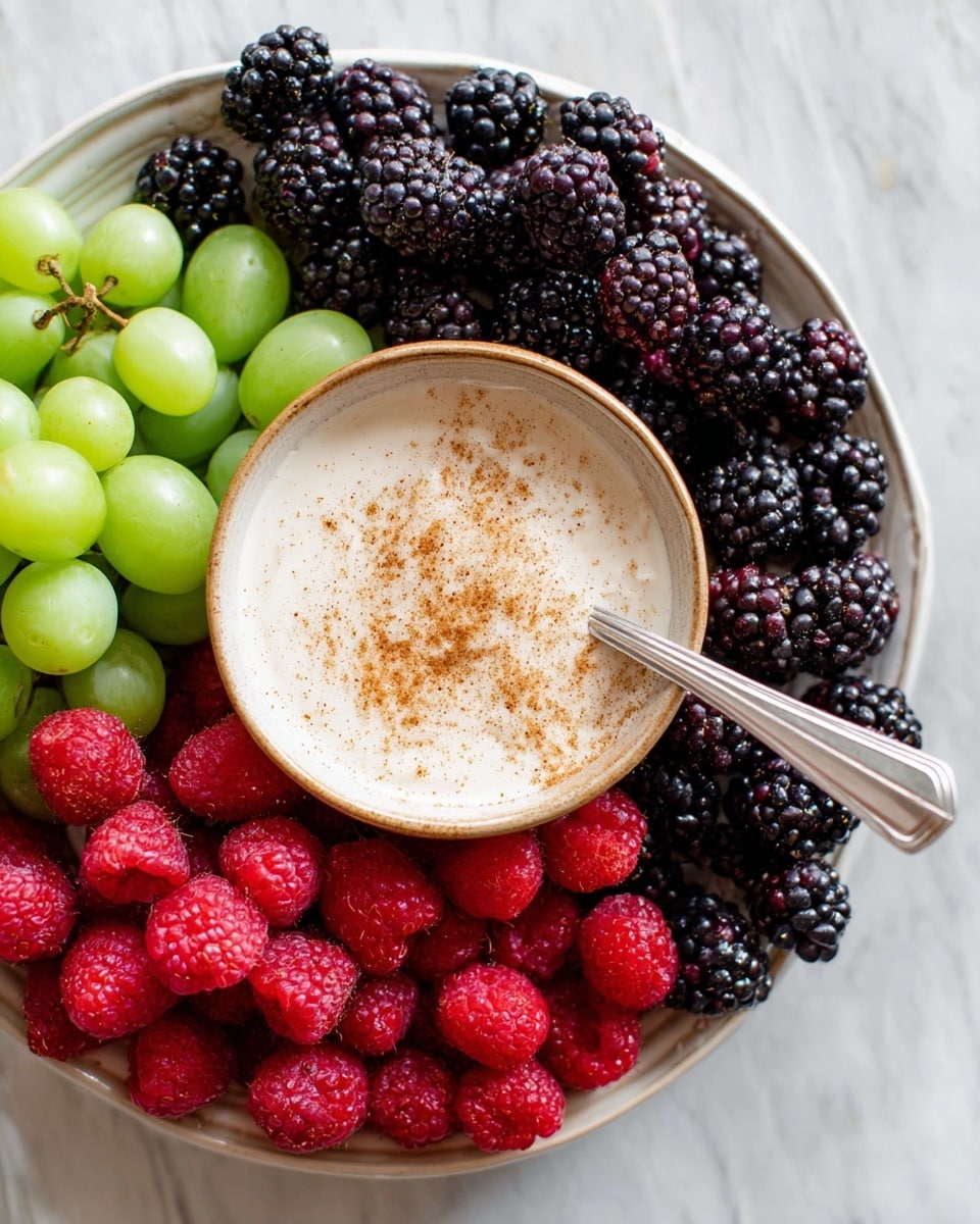 A round white plate filled with three layers of fresh fruit placed side by side: green grapes at the top left, blackberries in the middle and bottom left, and bright red raspberries on the right. In the center of the plate, there is a small beige bowl filled with creamy white dip topped with a light sprinkle of brown spice. A silver spoon rests inside the bowl, extending out towards the raspberries. The plate is set on a white marbled surface. Photo taken with an iphone --ar 4:5 --v 7